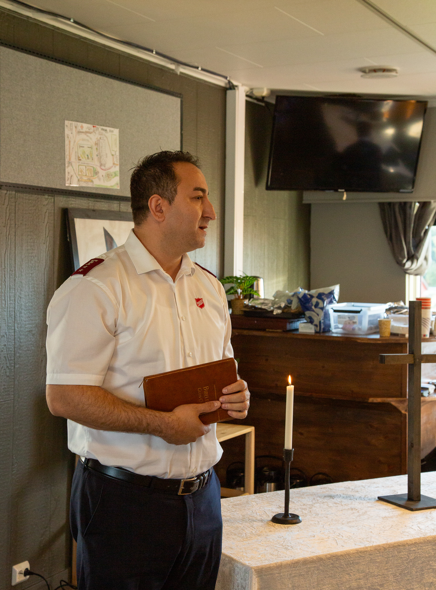 Foto: Mann i 50-årene i hvit uniformsskorte fra Frelsesarmeen og mørke bukser står ved et bord med pen duk. Han holder en bibel  under høyre arm. Ved siden av ham står et tent stearinlys.