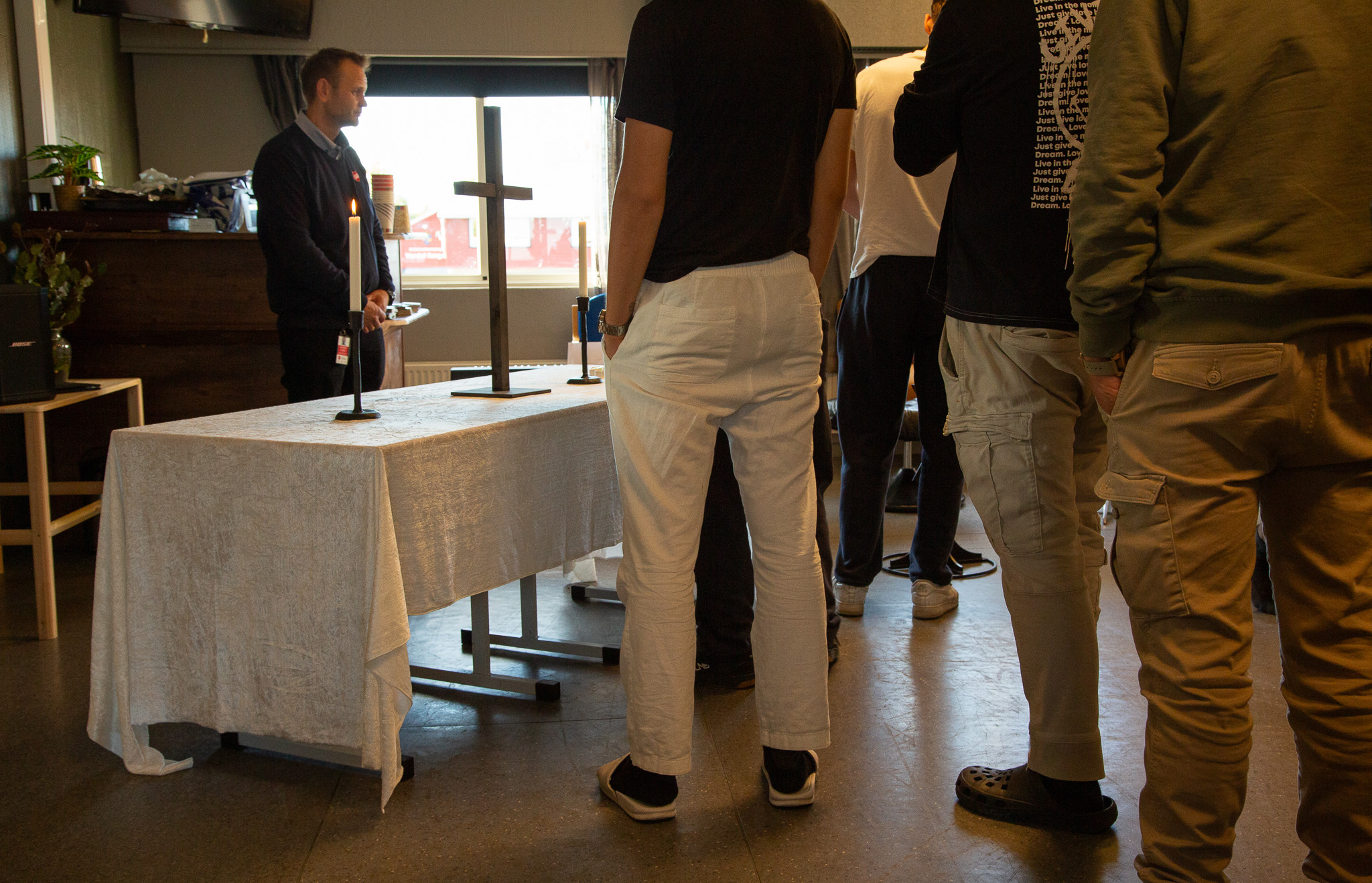 Foto: Menn som står i kø inne i et rom. Et bord med duk og et trekors på, bak dem er det et vindu ut mot en gressplen.