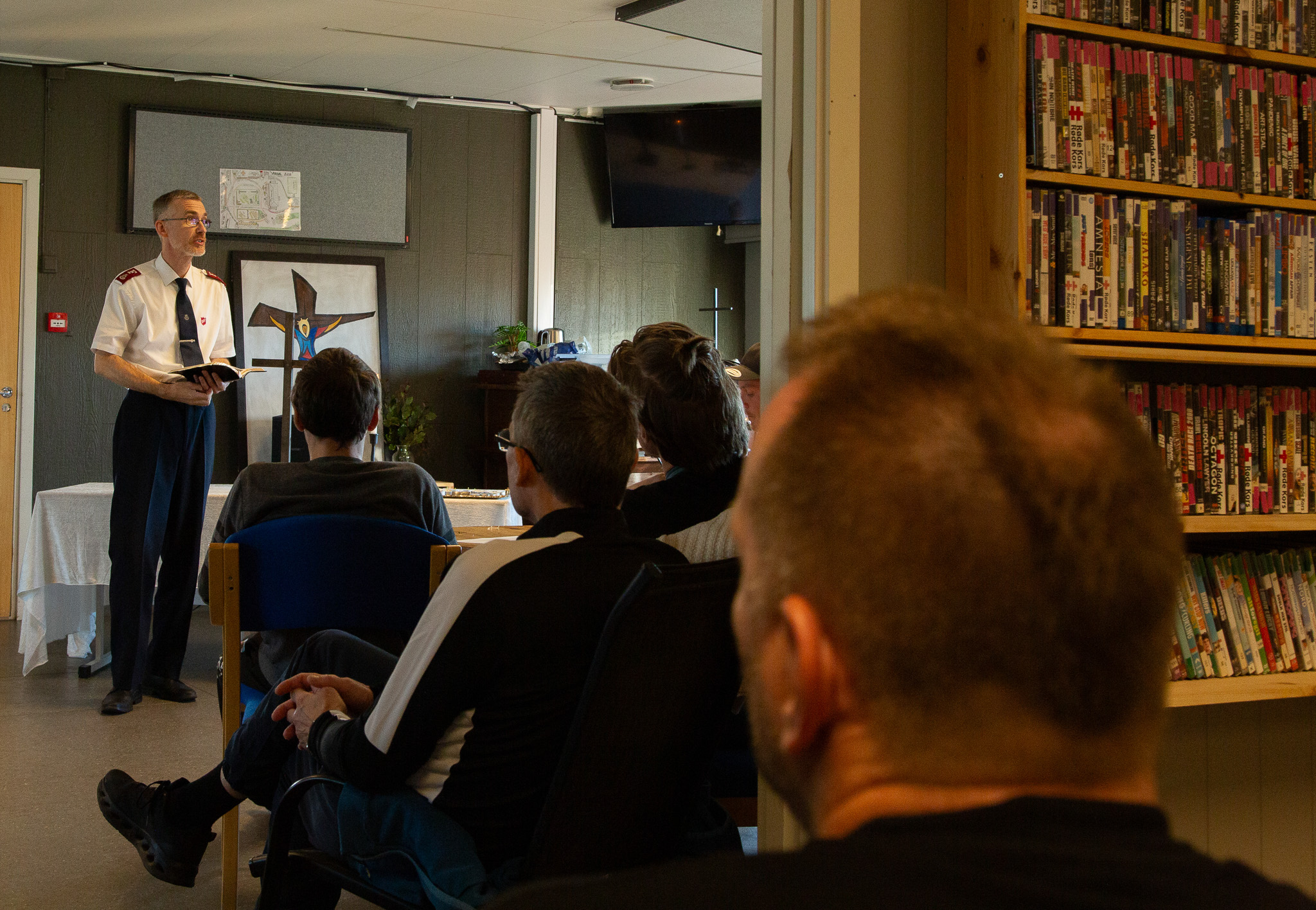 Inne i et rom står en mann i uniform med en bibel i hendene. Foran ham sitter flere menn på stoler.