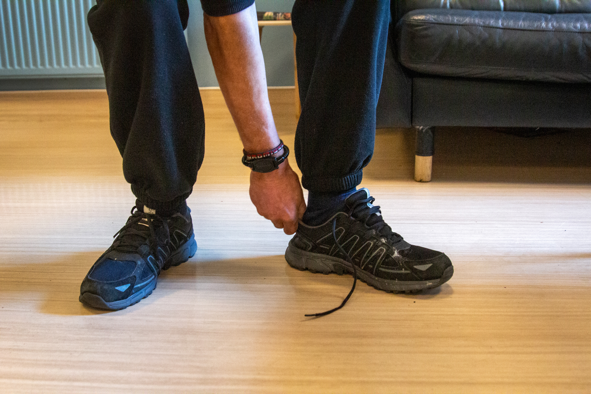 Nærbilde av beina til en mann som har svart jobbebukse og knytter på seg svarte joggesko.