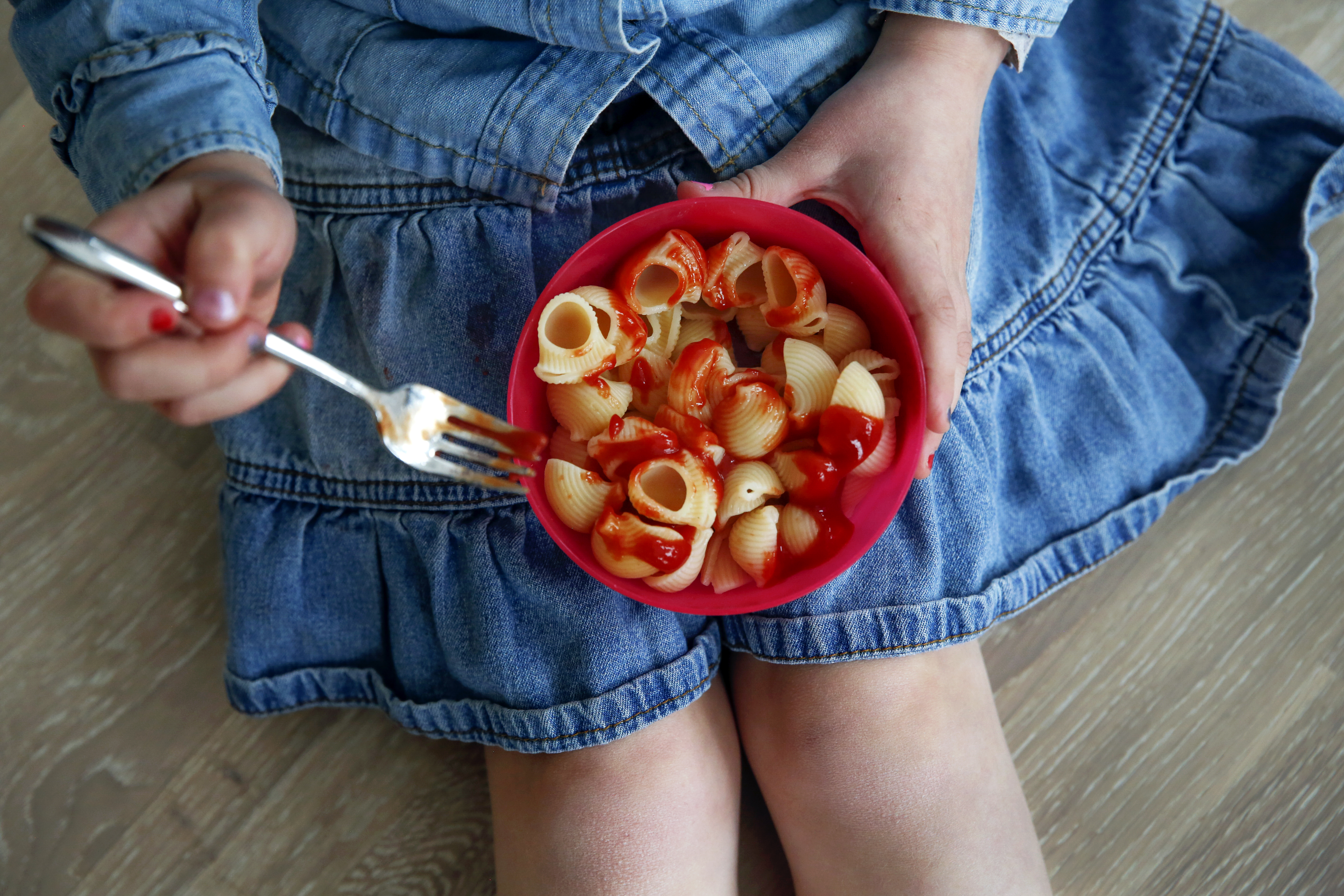 Barn som sitter med en plastskål med spaghetti og ketchup. Ser bare hender og fanget til barnet.