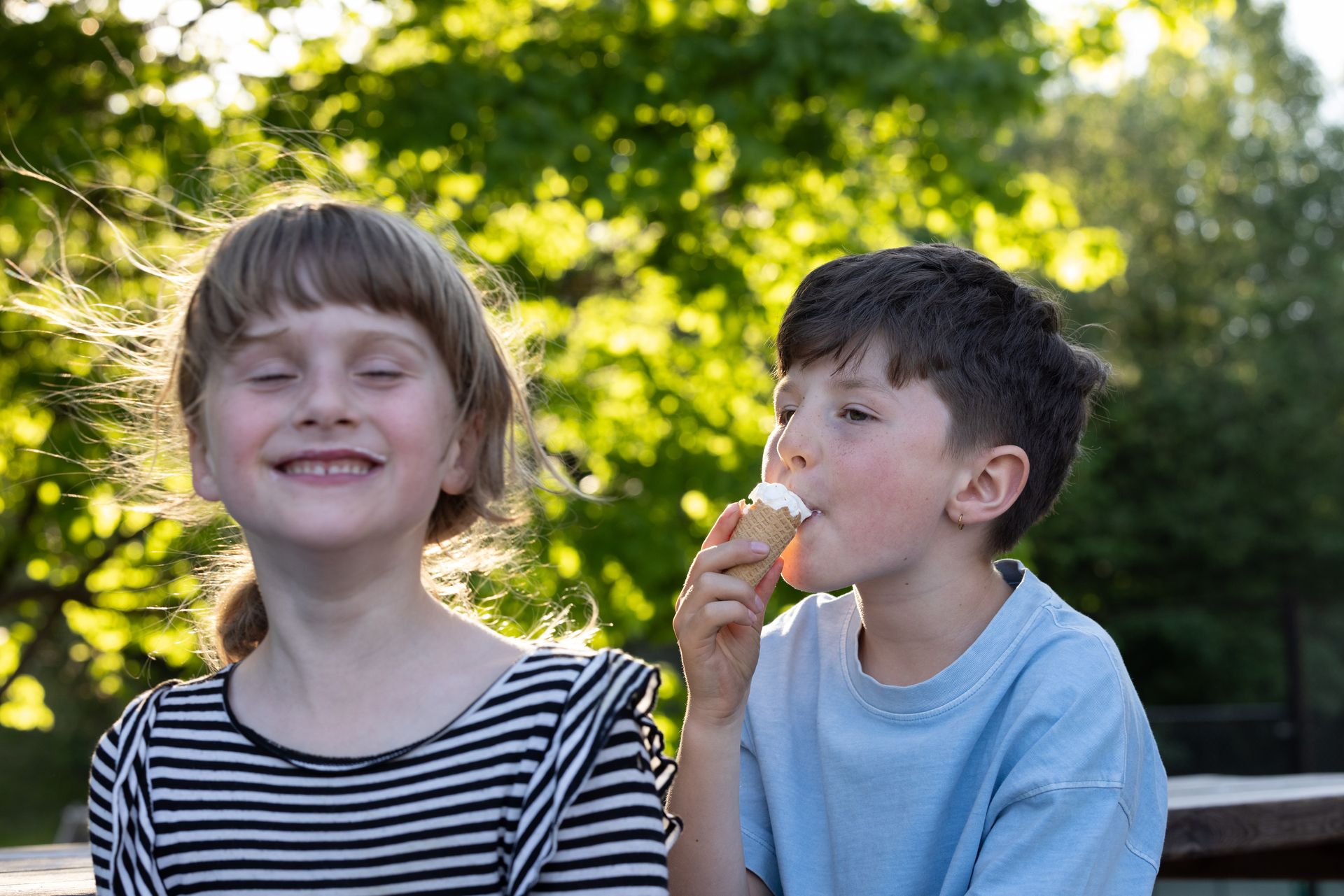 En jente på rundt 9 år ser i kameraet og gliser bredt med litt rester av is rundt munnen. Vi ser en gutt på cirka 12 fra siden fornøyd spise en kronesis.