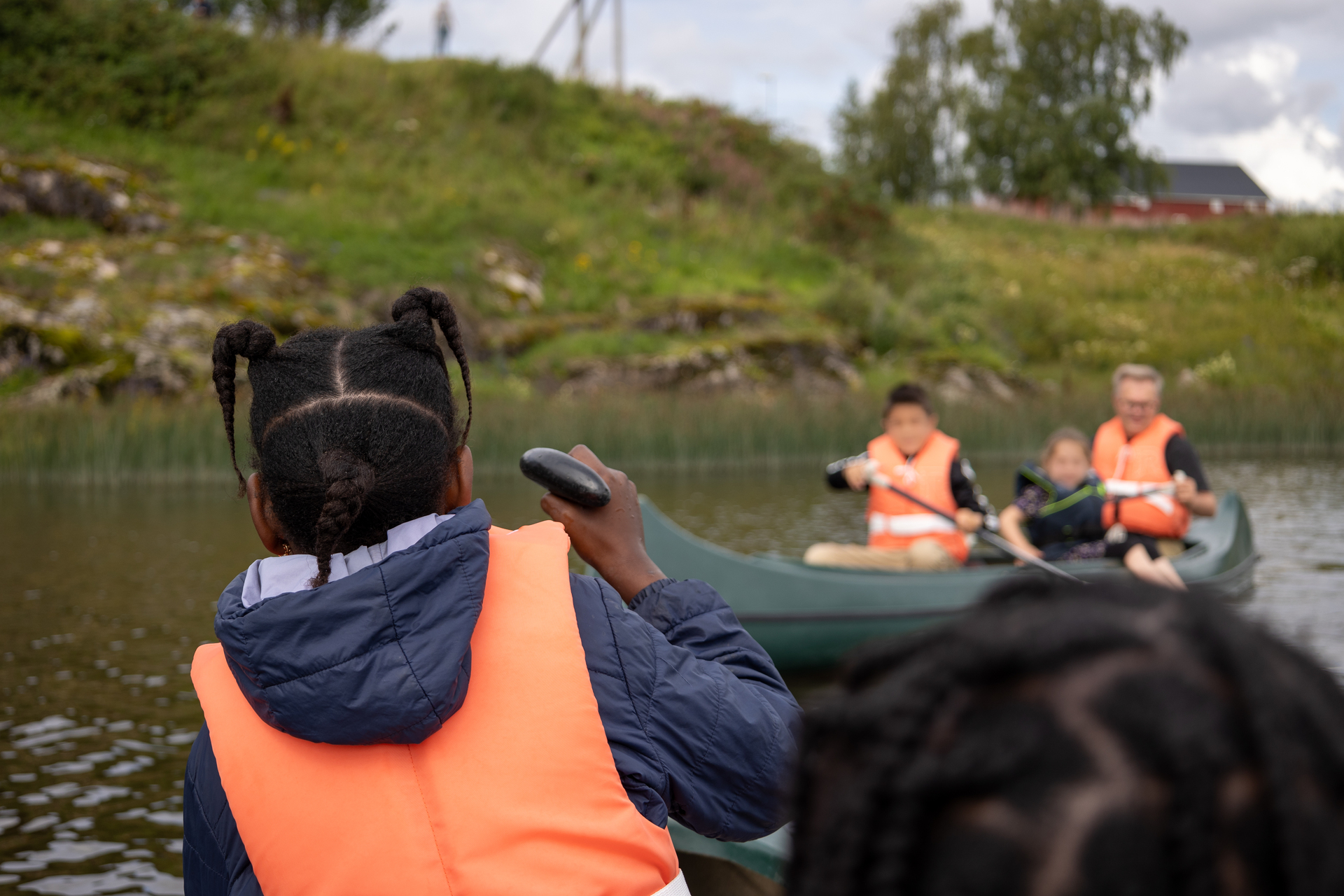 ¨Vi ser bakhodet til en jente med flytevest. Hun ser mot en kano på vannet med tre personer. Alle med redningsvest. en voksen leirleder sitter i kanoen.