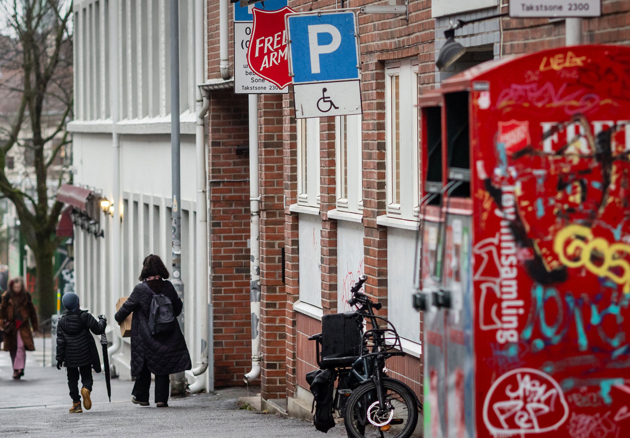 Foto: En dame og en gutt går nedover en gate. Hun bærer på en eske. På en murbygning bak dem henger et skilt fra Frelsesarmeen. Og helt i forgrunnen av bildet står et par nedtaggede innsamlingsbokser fra Fretex.