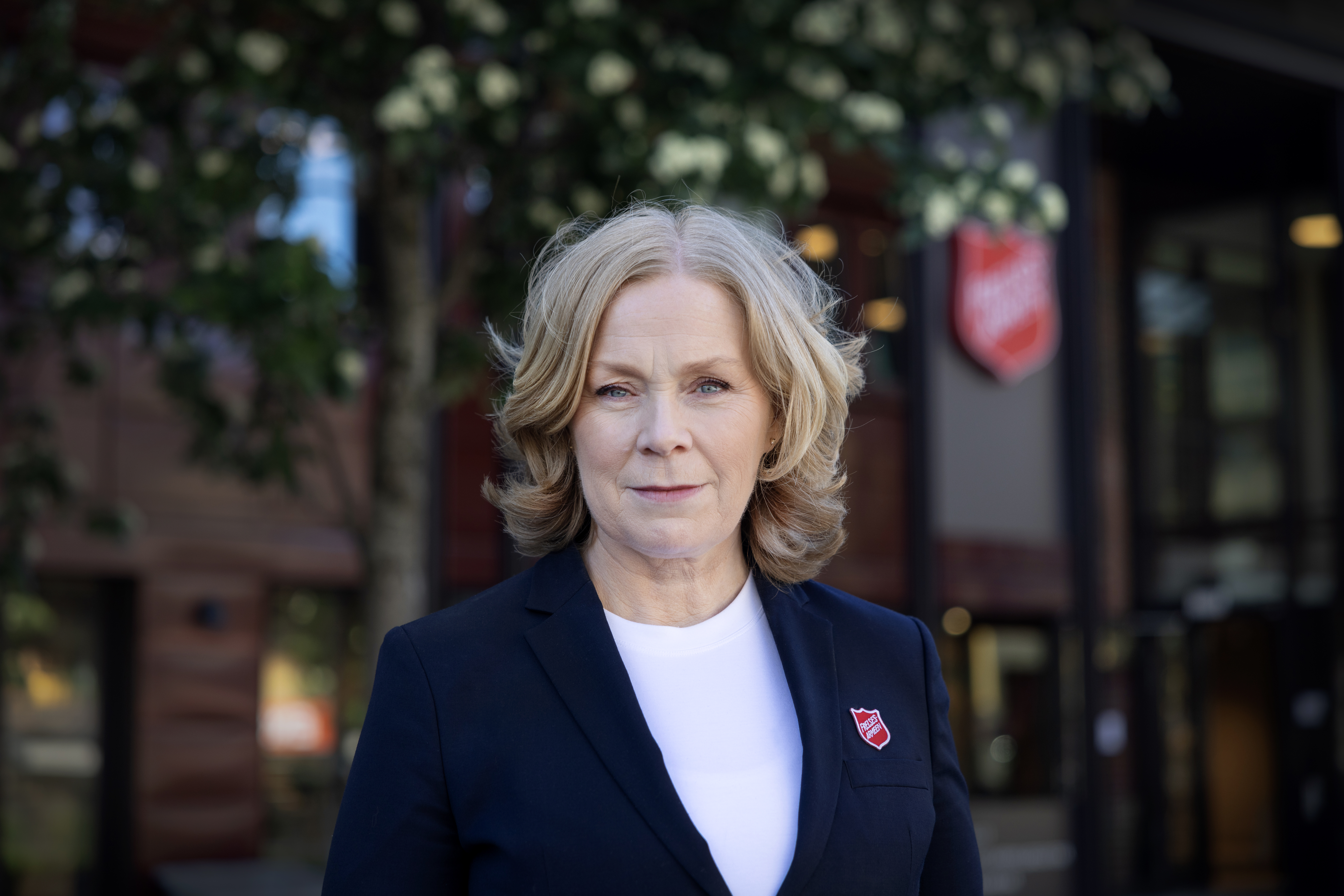 Foto: bilde av alvorlig dame i Frelsesarmeen-uniform, fra midjen og opp, med uklar bakgrunn med trær og et bygg.