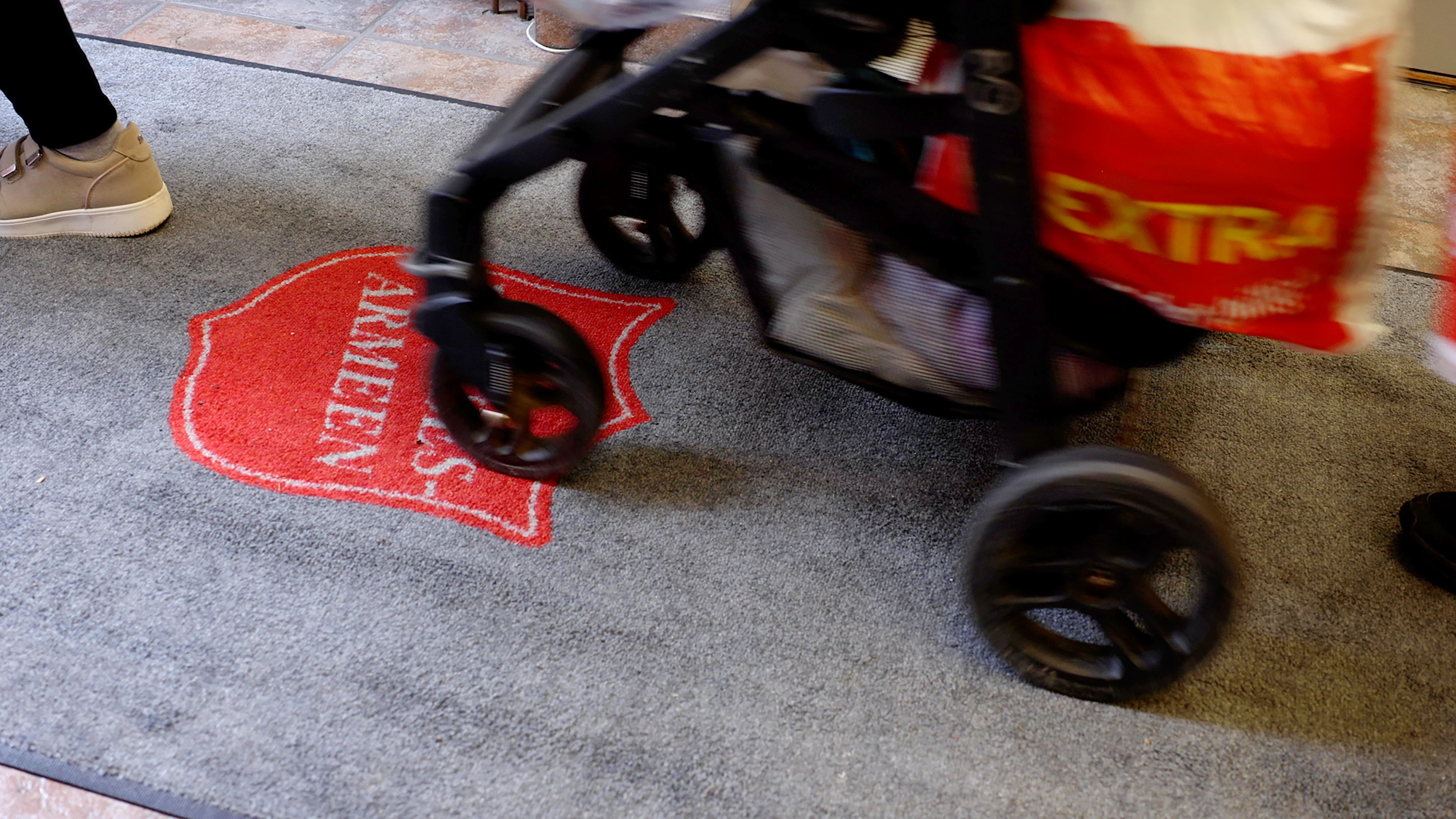 Foto: En barnevogn som ruller ever en grå løper med skjoldet til Frelsesarmeen på. På vogna henger det en pose med matvarer. Vi ser ingen mennesker, kun føtter med sko.