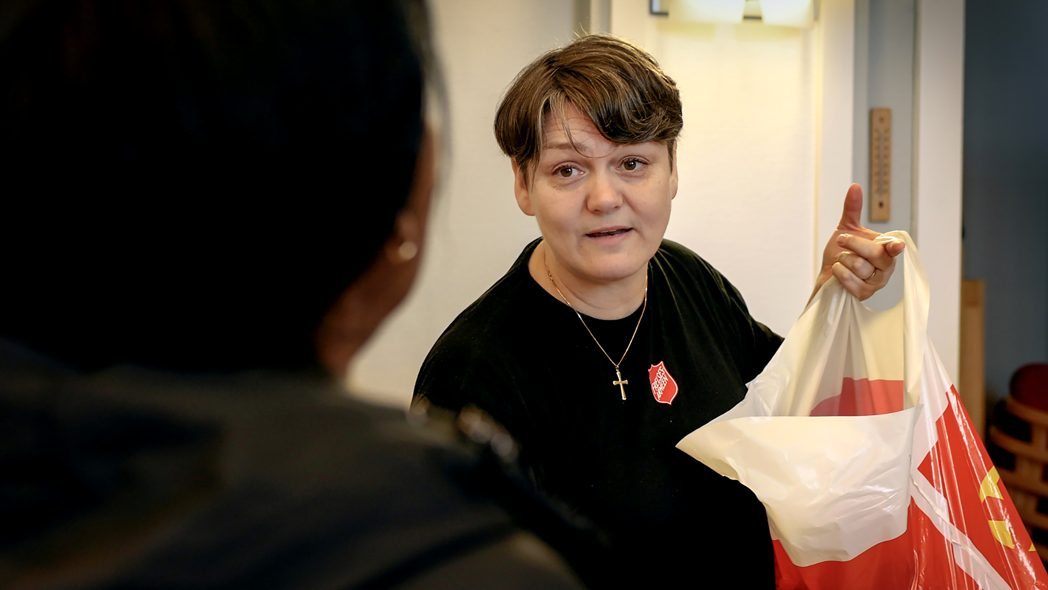 Foto: Kvinne sent i 40-årene med logo fra Frelsesarmeen på genseren og gullkors rundt halsen, holder opp en pose med matvarer. Hun ser på noen vi ser ryggen til.