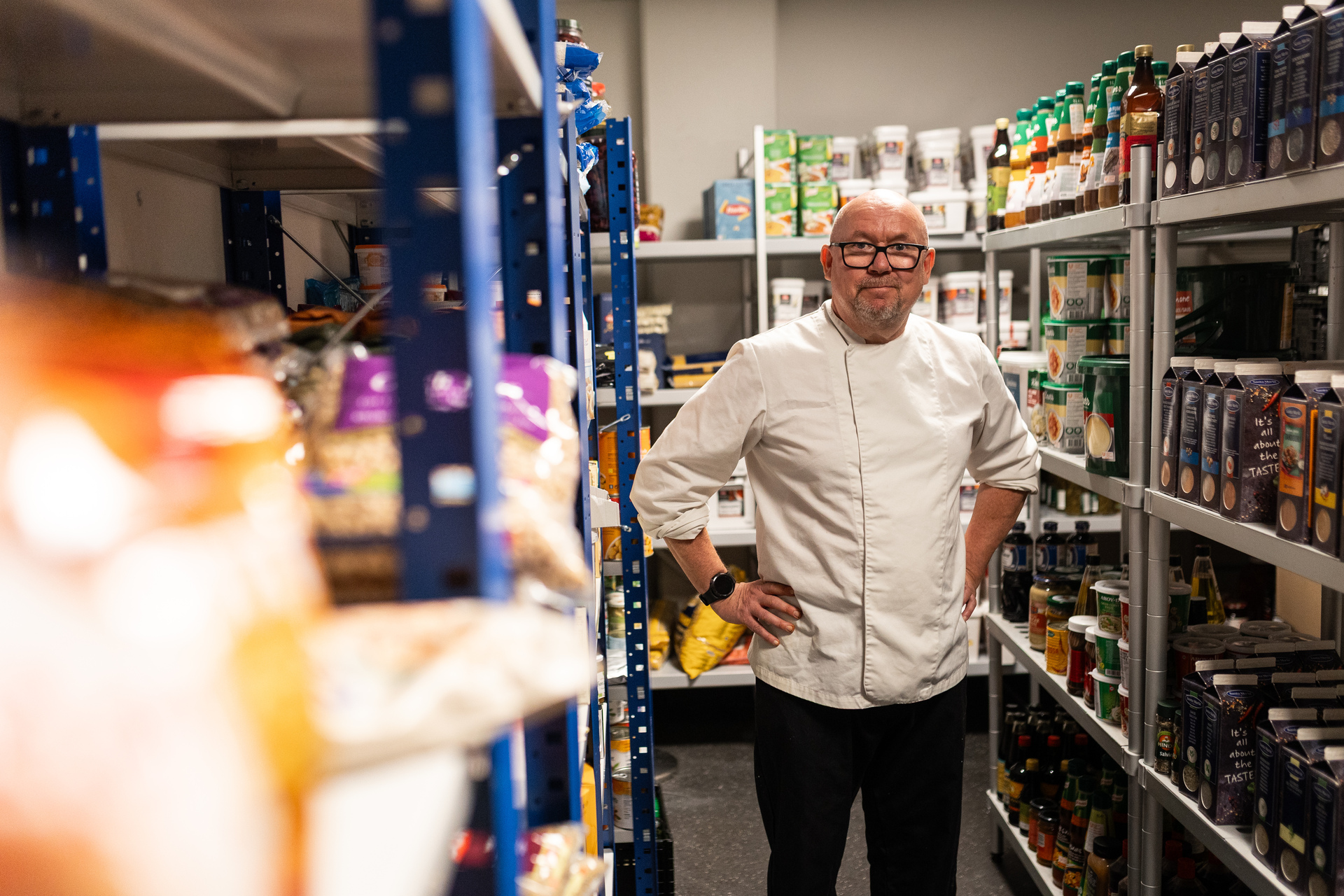 Foto: En skallet mann med hvit kokkejakke og svarte bukser står med hendene i siden og ser mot kamera. Han står på et lager mellom hyller med tørrvarer og sauser.