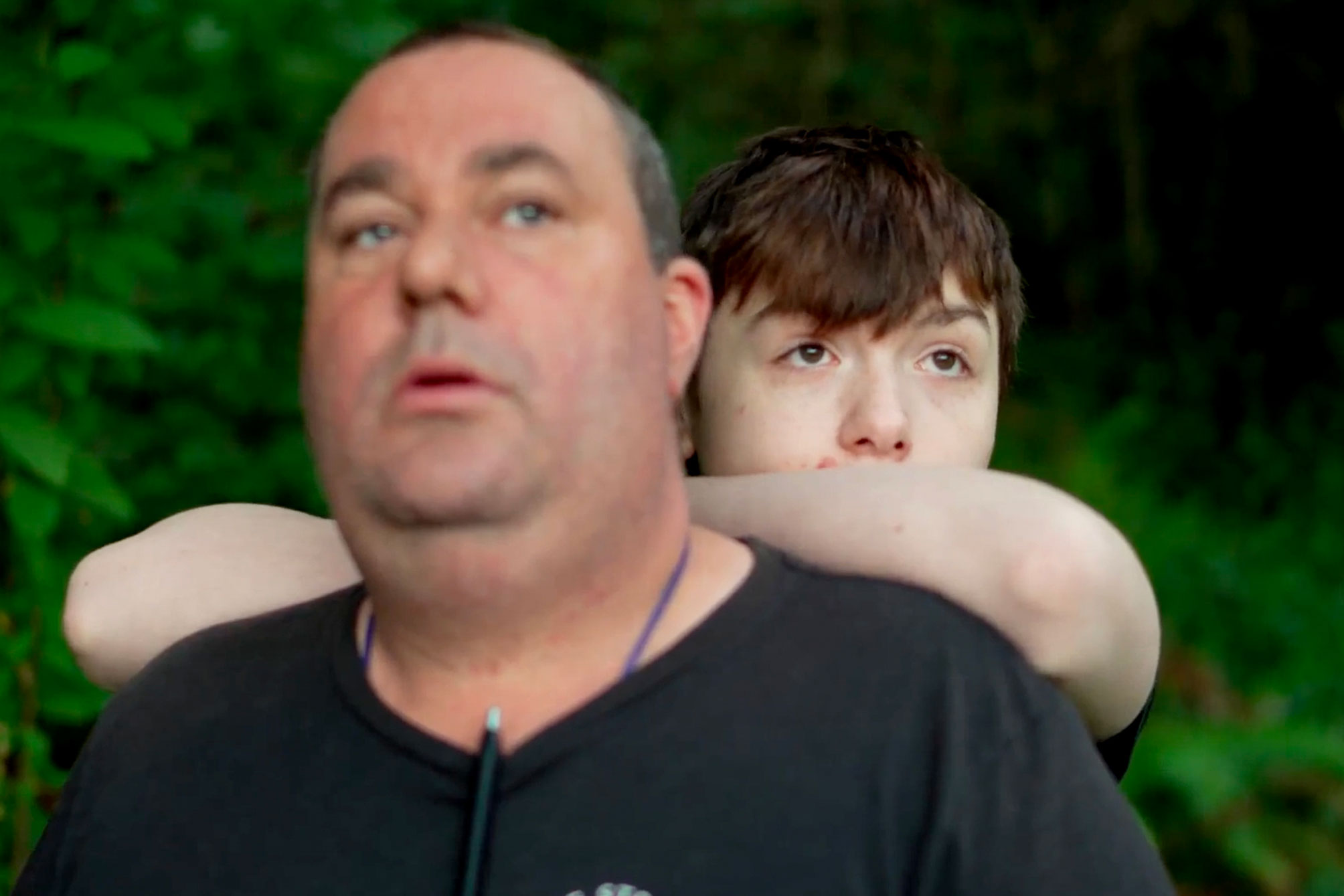 Foto: Gutt står bak faren sin. Han lener seg på ryggen hans. Begge ser alvorlige ut