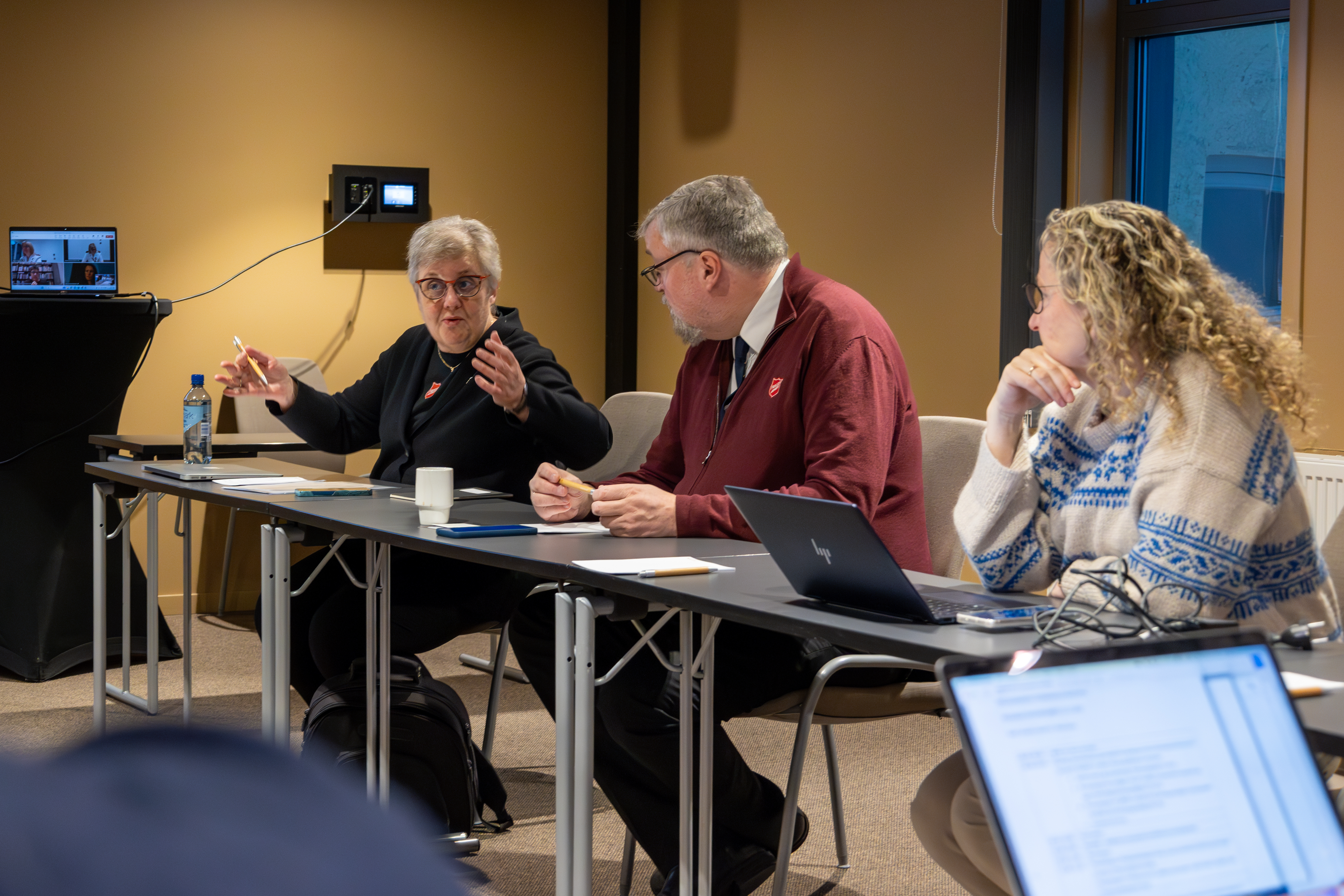 Tre sitter på en rekke, der kvinnen som snakker gestikulerer med hendene.