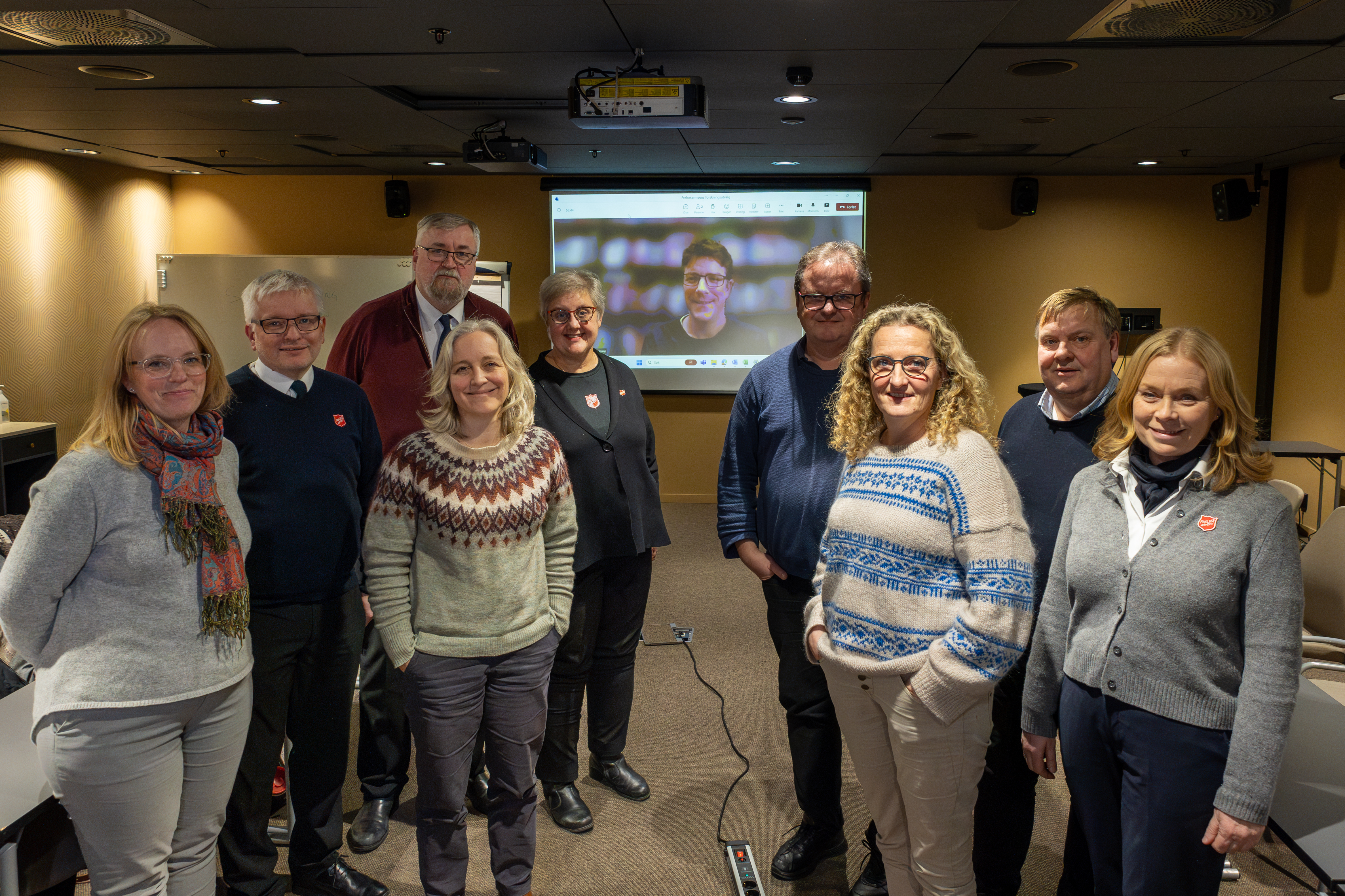 Ni personer står oppstilt foran en skjerm på veggen der du ser en mann.