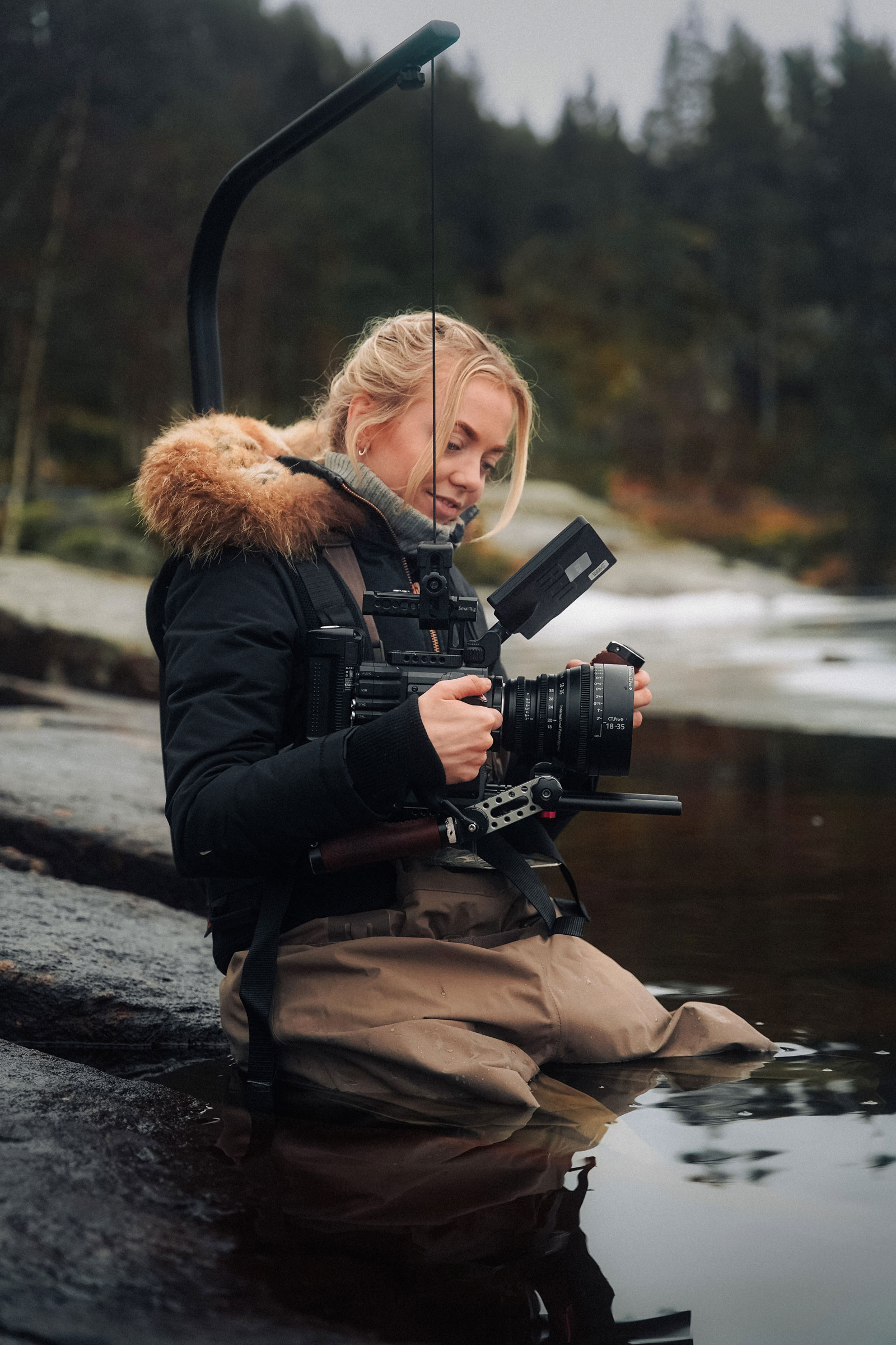 Foto: Kvinne 20-årene med blondt hår holder et videokamera med begge hendene. Hun sitter på et svaberg, halvvegs ut i vannet. Hun har på seg beige vadebukser og en svart boblejakke med pelskrage.