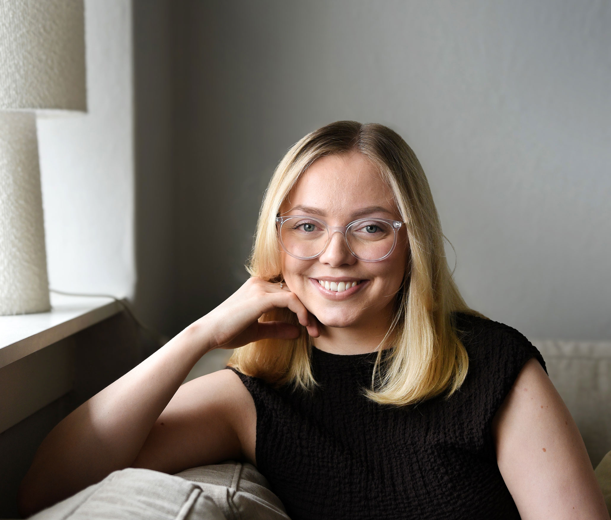 Foto: Blond kvinne med blondt hår nedpå skuldrene smiler mot kamera. Hun har på seg briller med lys, nesten gjennomsiktig kant og smiler så store, hvite tenner viser. Hun er kledd i svart og støtter hodet mot høyre arm oppå ryggputen i sofaen.
