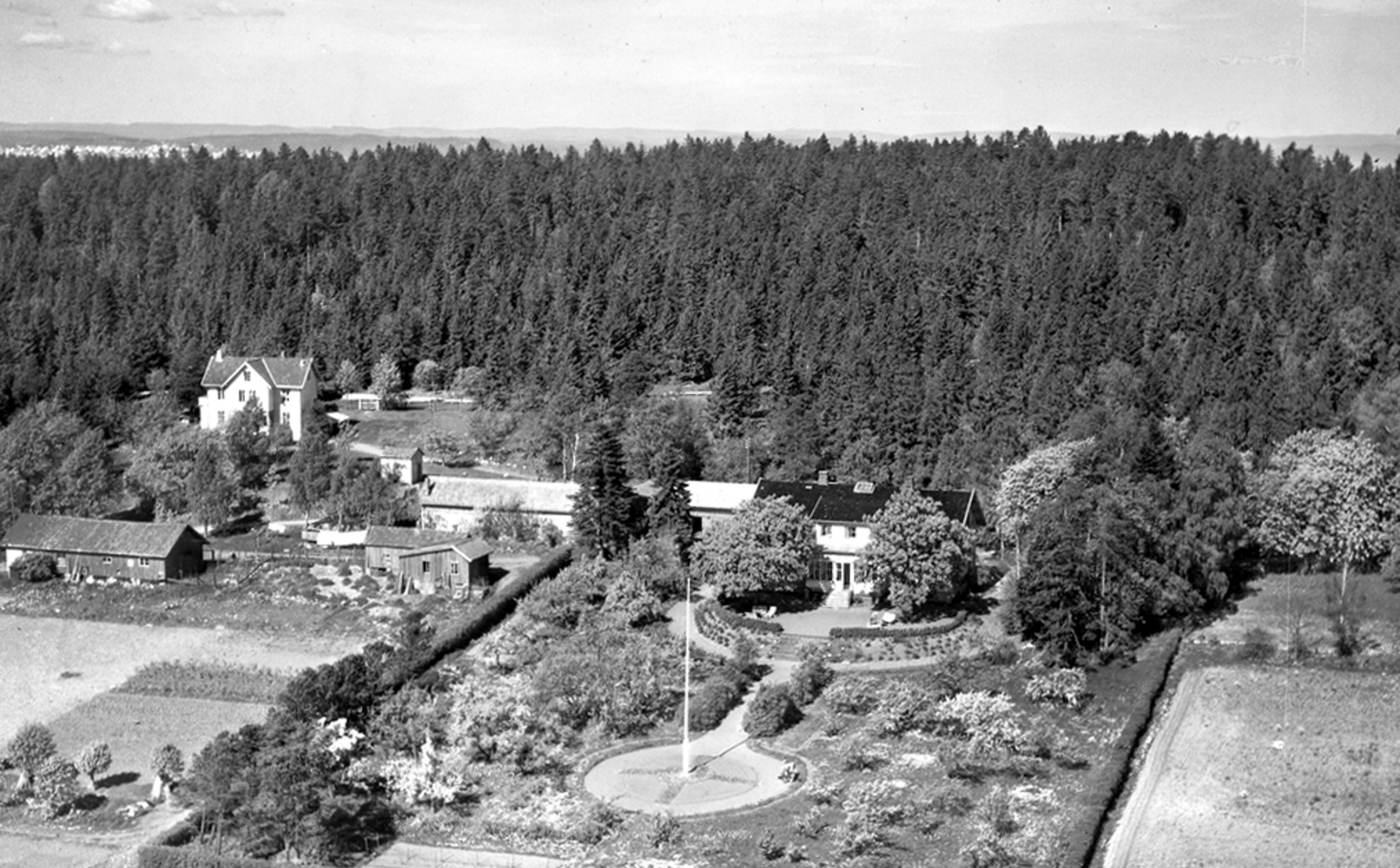 Svart hvitt bilde av et gammelt murhus med en parklignenede hage i front. Bak ligger en skog, noen mindre uthus og et  stort toetasjes hus i tre.