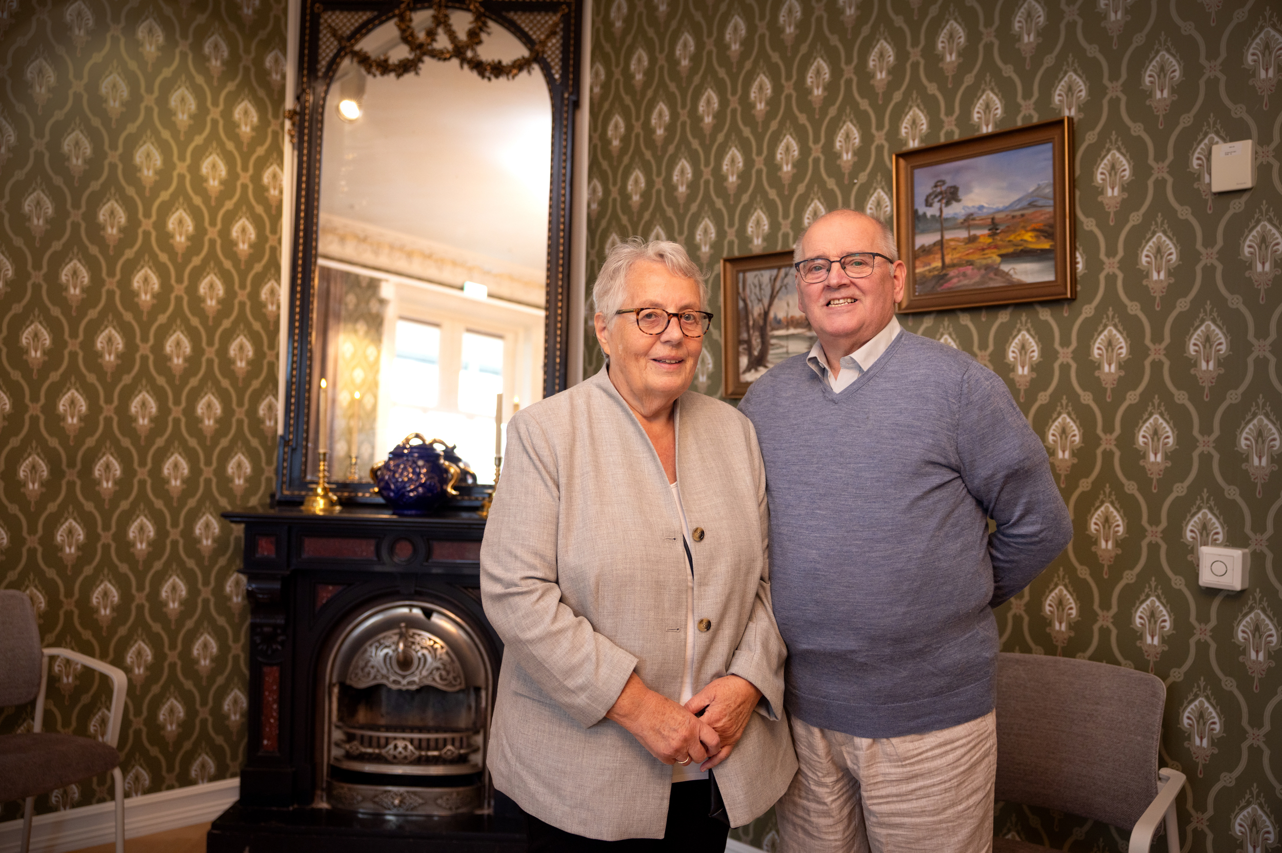 Dame og mann i slutten av 70-årene står pent oppstilt ved siden av hverandre foran en gammel vedovn med et speil oppå. På veggene er det en grønn tapet med mønster i hvitt og gull.