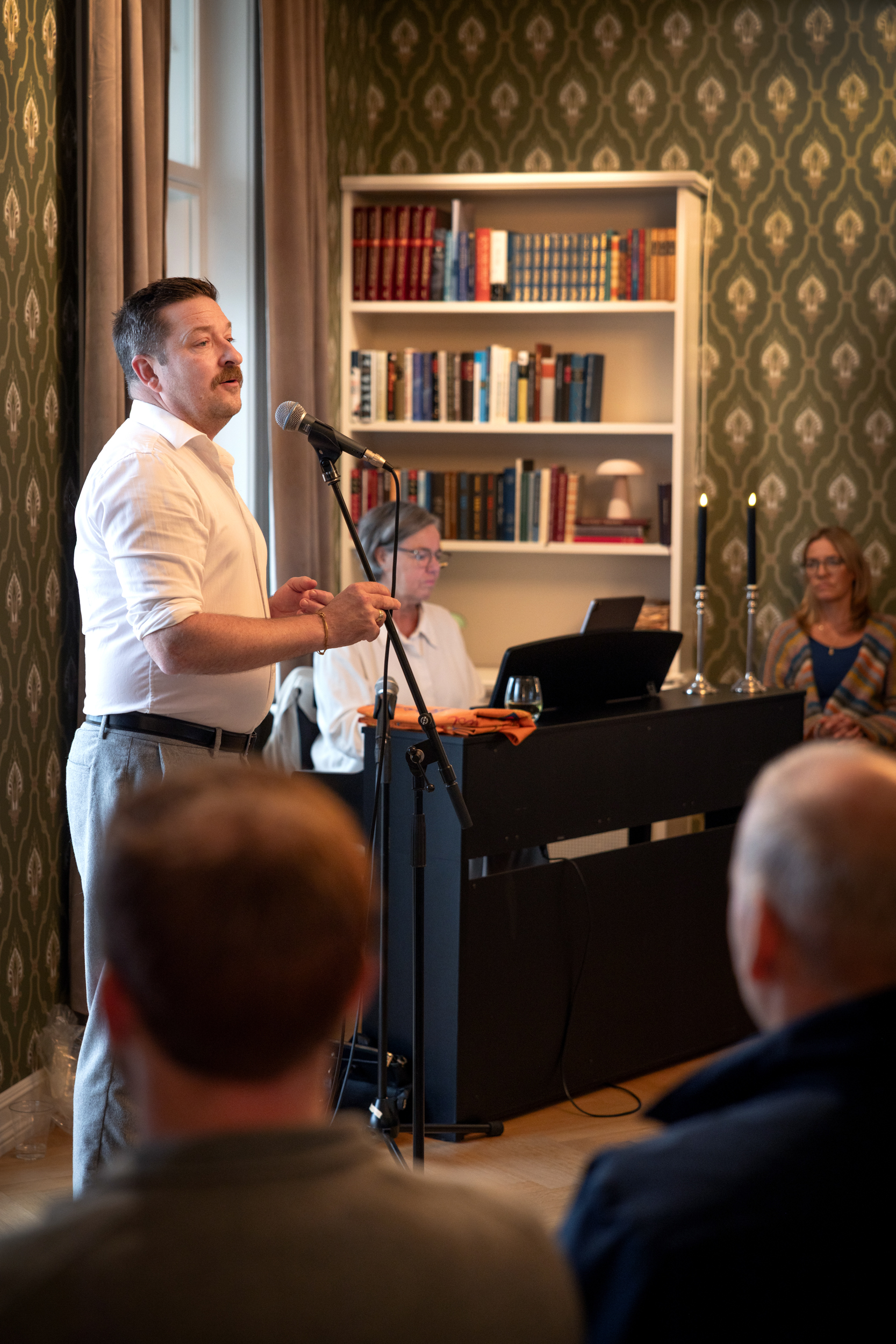 Mann med mørkt hår og bart synger i et mikrofonstativ. I bakgrunnen sitter det en kvinne på piano. Hun har grått hår og briller. Det er bøker i en bokhylle på veggen bak og folk som sitter og ser på.