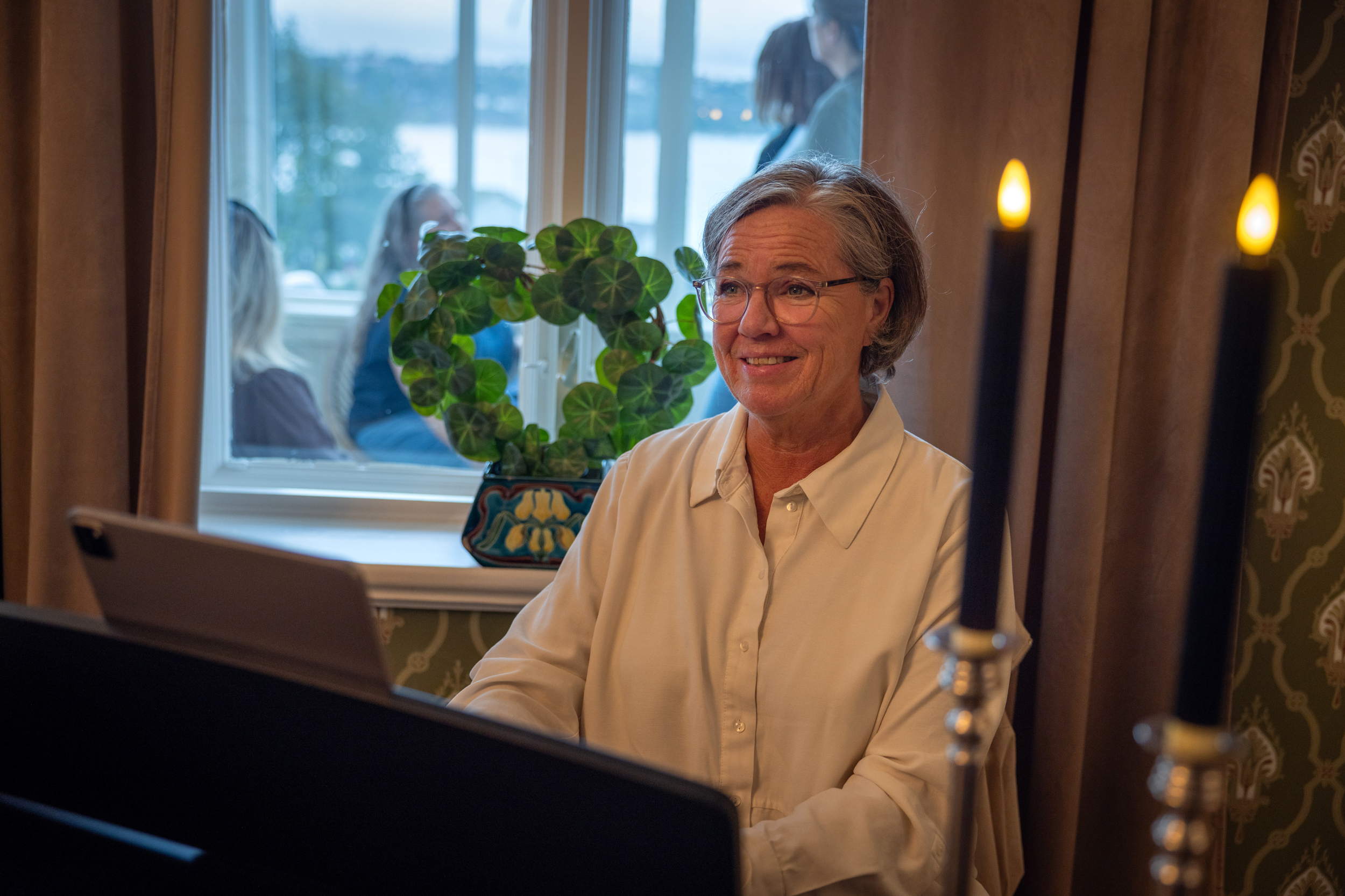 Kvinne tidlig i 60-årene sitter ved et piano og spiller. Hun har kort, grått hår og briller.