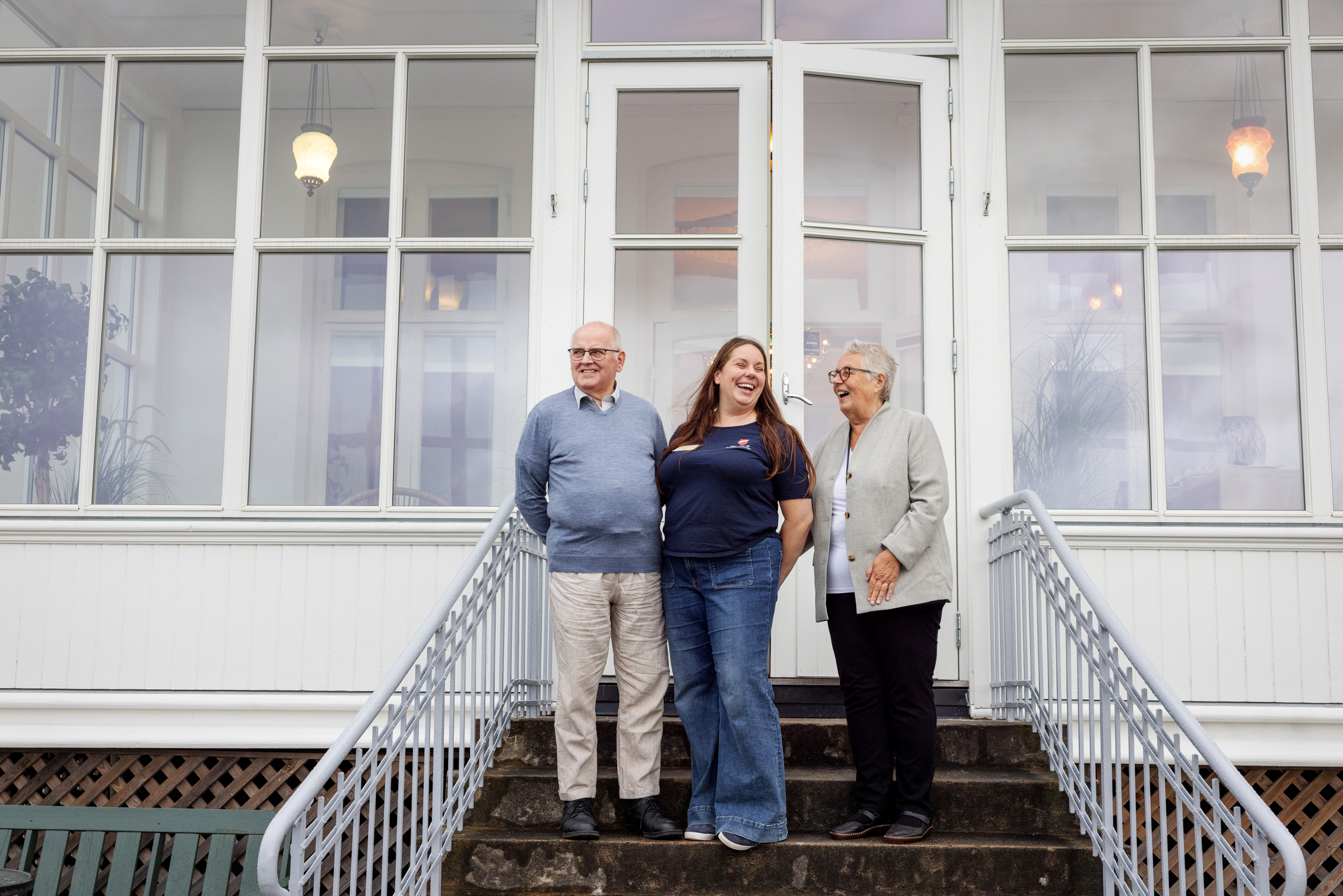 Kvinne og mann i 70-årene står sammen med en kvinne i 40-årene med langt, brunt hår. De str på en trapp med rekkverk opp til en vinterhage i hvitt tre og glass.