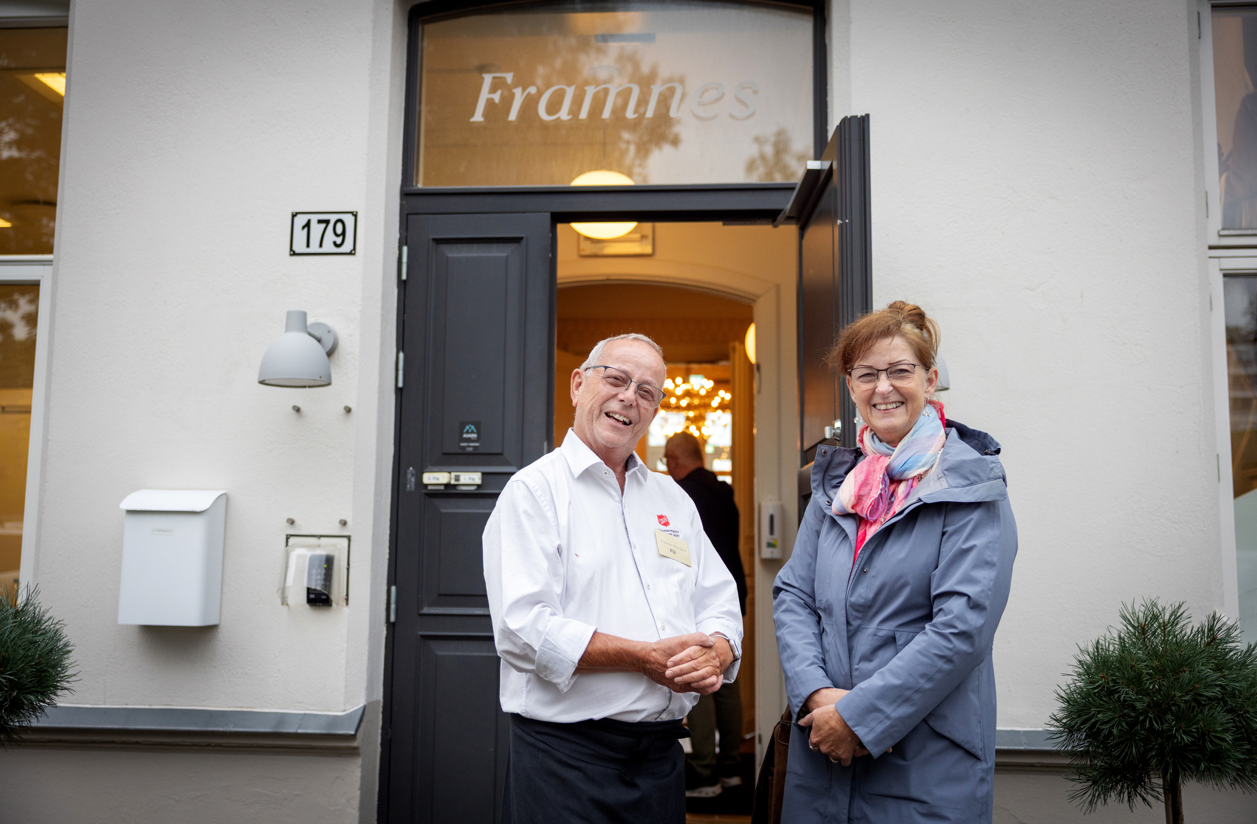 Mann i 60-årene med briller og i hvit skjorte med logo fra Frelsesarmeen hilser på en voksen dame med mørkt, oppsatt hår og briller. De står utenfor et hvitt murhus.