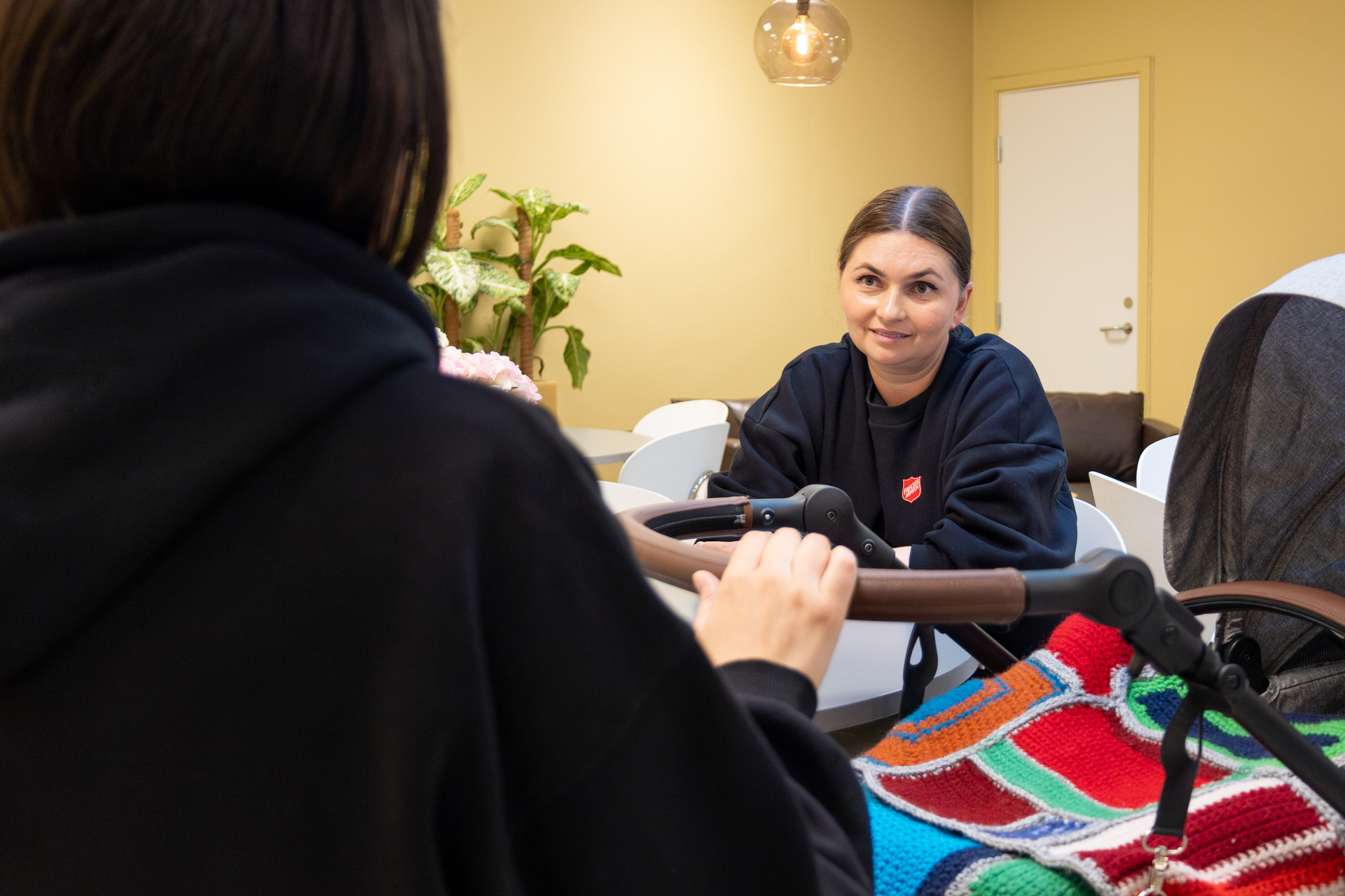 Foto: Kvinne som holder i en barnevogn sitter med ryggen til. Hun snakker med en ansatt hos Frelsesarmeen. De to kvinnene sitter ved et bord og snakker.