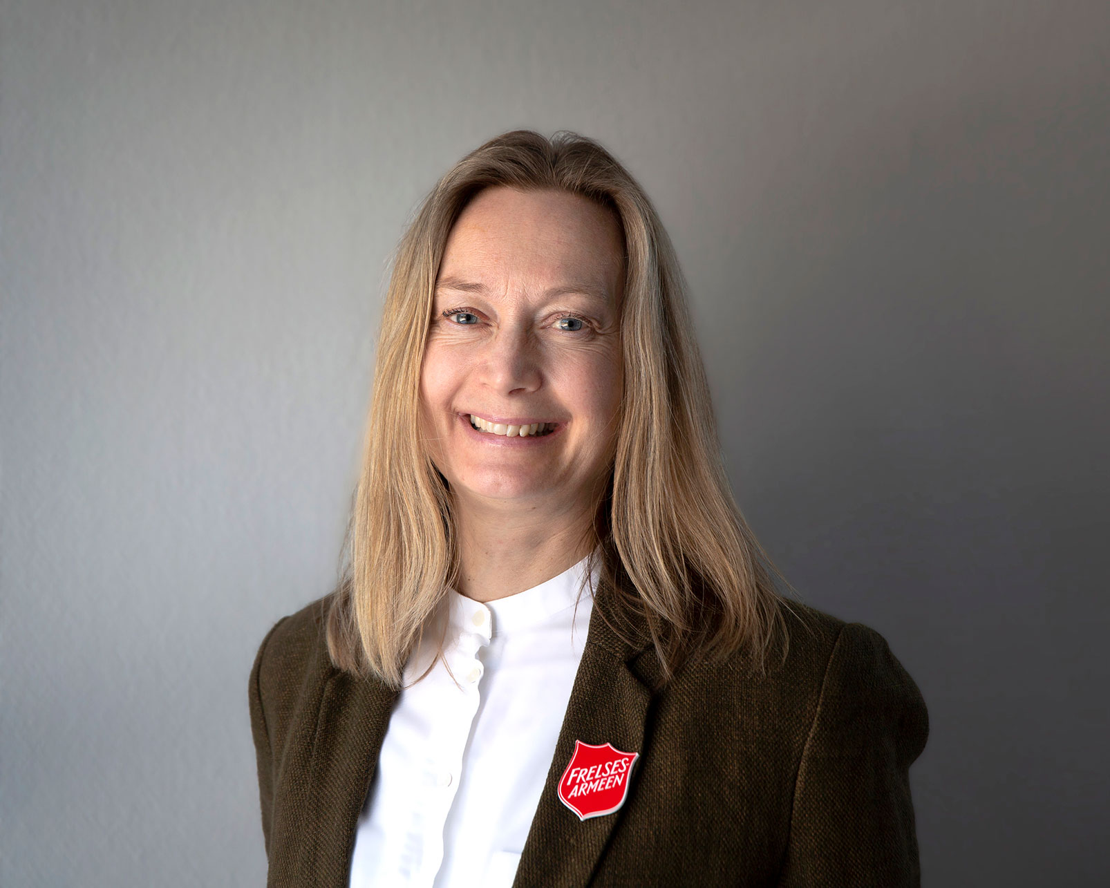 Foto: Portrettbilde av kvinne i 40-årene med langt lyst hår. Hun har på seg hvis bluse og blå dressjakke med rød logo fra Frelsesarmeen.
