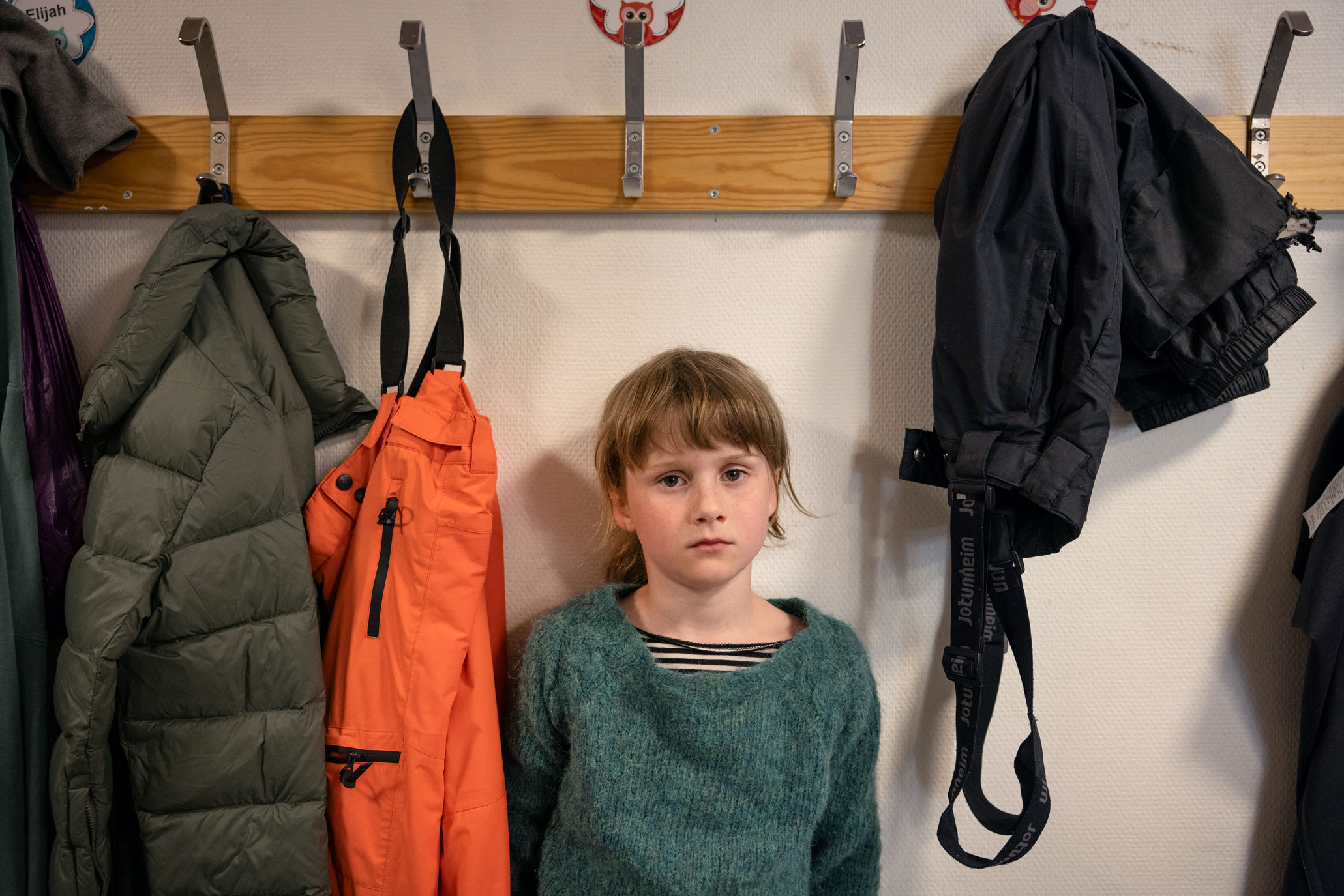 Foto: Jente rundt 10 år med blondt hår står i en garderobe. Her henger det klær på knaggene og hun ser trist ut.