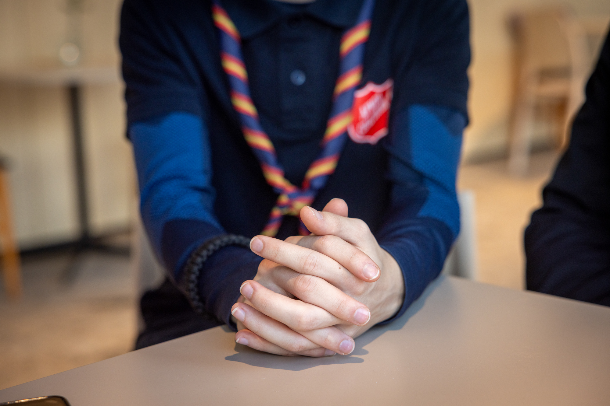 Foto: Unge guttehender som er foldet ligger på et bord. Personen som ber har på seg en mørkeblå genser med logo fra Frelsesarmeen og et knyttet, tvinnet skjerf i fargene gult, rødt og blått.