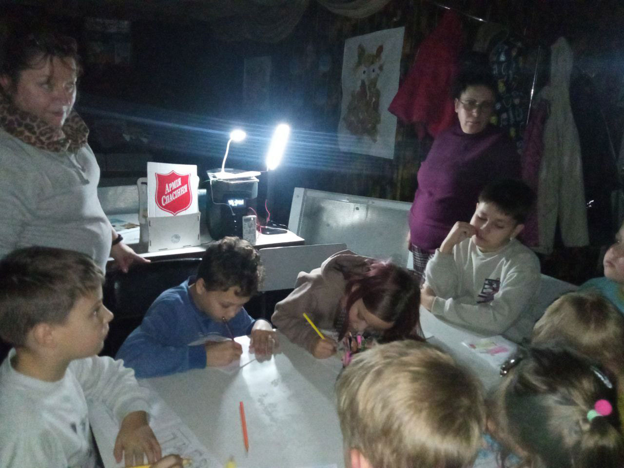Foto: Åtte barn sitter rundt et bord i et mørkt rom, det er kun to sterke lamper som lyser opp rommet fra et høyere bord. Ved siden av står to voksne damer og mellom dem står det et skjold fra Frelsesarmeen.