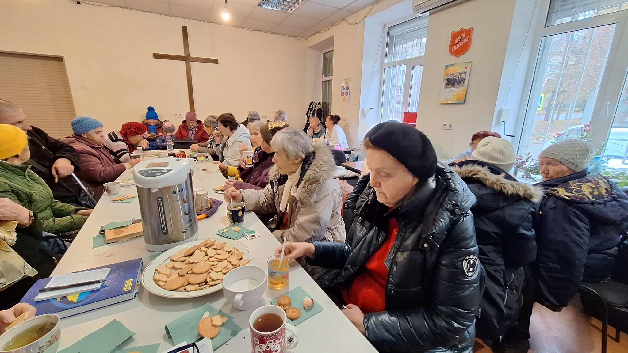 Foto: Mange eldre mennesker sitter påkledde i et møterom innendørs. På den ene veggen er det et stort kors og på den andre et rødt skjold der det står Frelsesarmeen på ukrainsk. På bordene står det fat med kjeks og store kanner med kaffe.