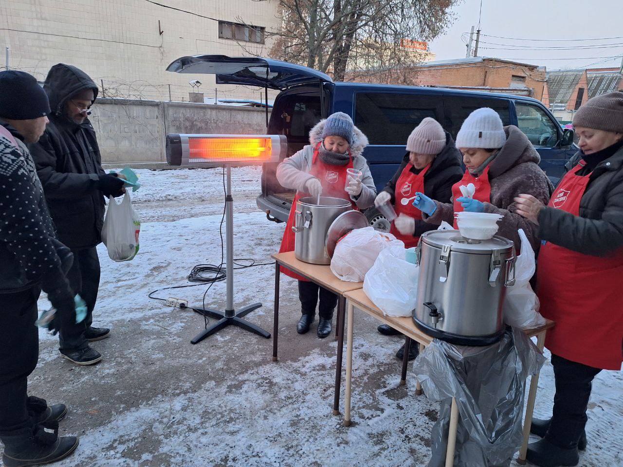 Foto: Fire damer i røde forklær med logo fra Frelsesarmeen står ute i snøen og serverer mat fra et bord til mennesker. Det står en varebil like bak som det kommer en ledning ut av og som gir strøm til en glødende varmelampe.