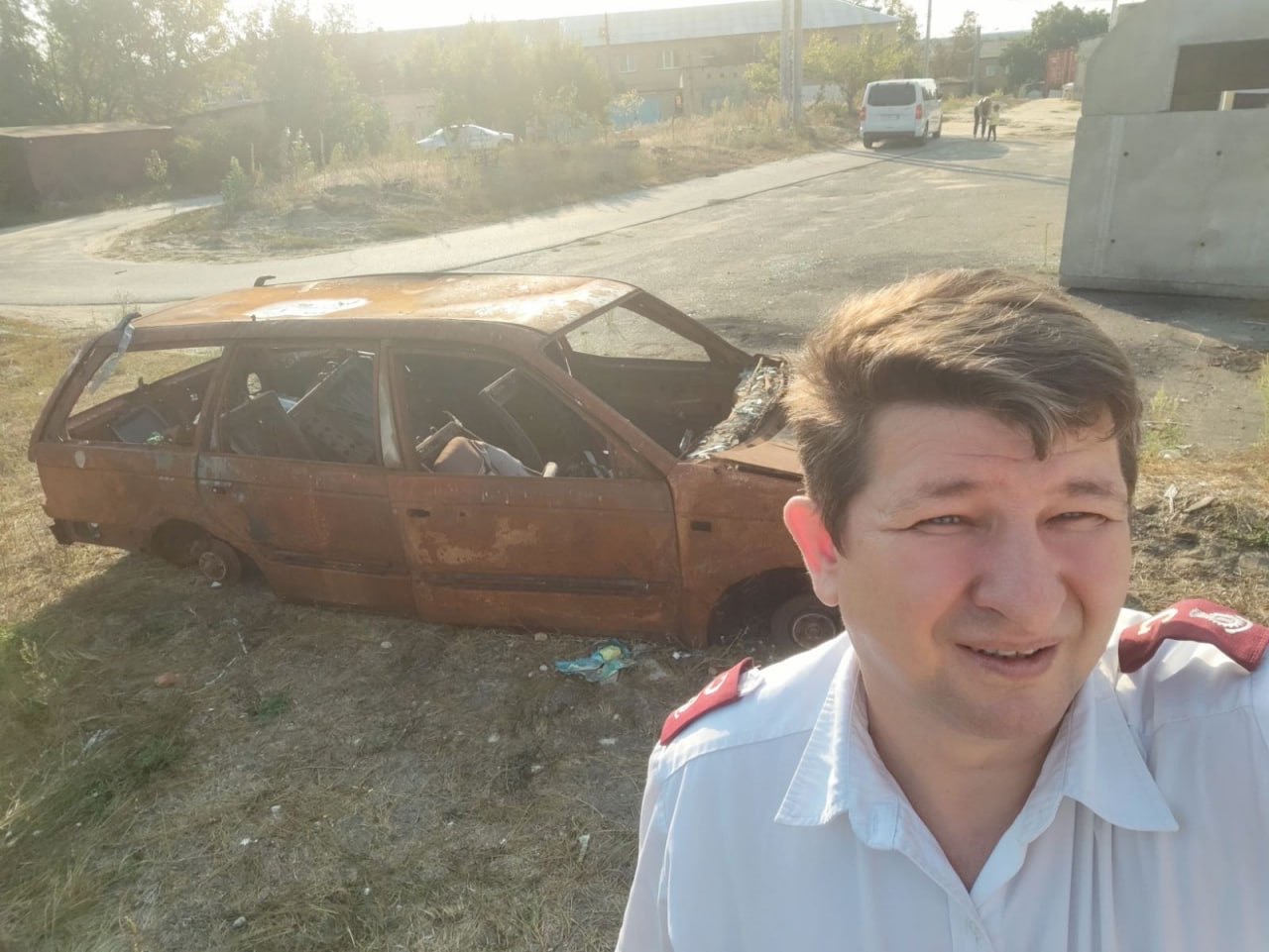 Foto: Mann i hvit skjorte fra Frelsesarmeen tar selfie med en totalt utbrent bil.