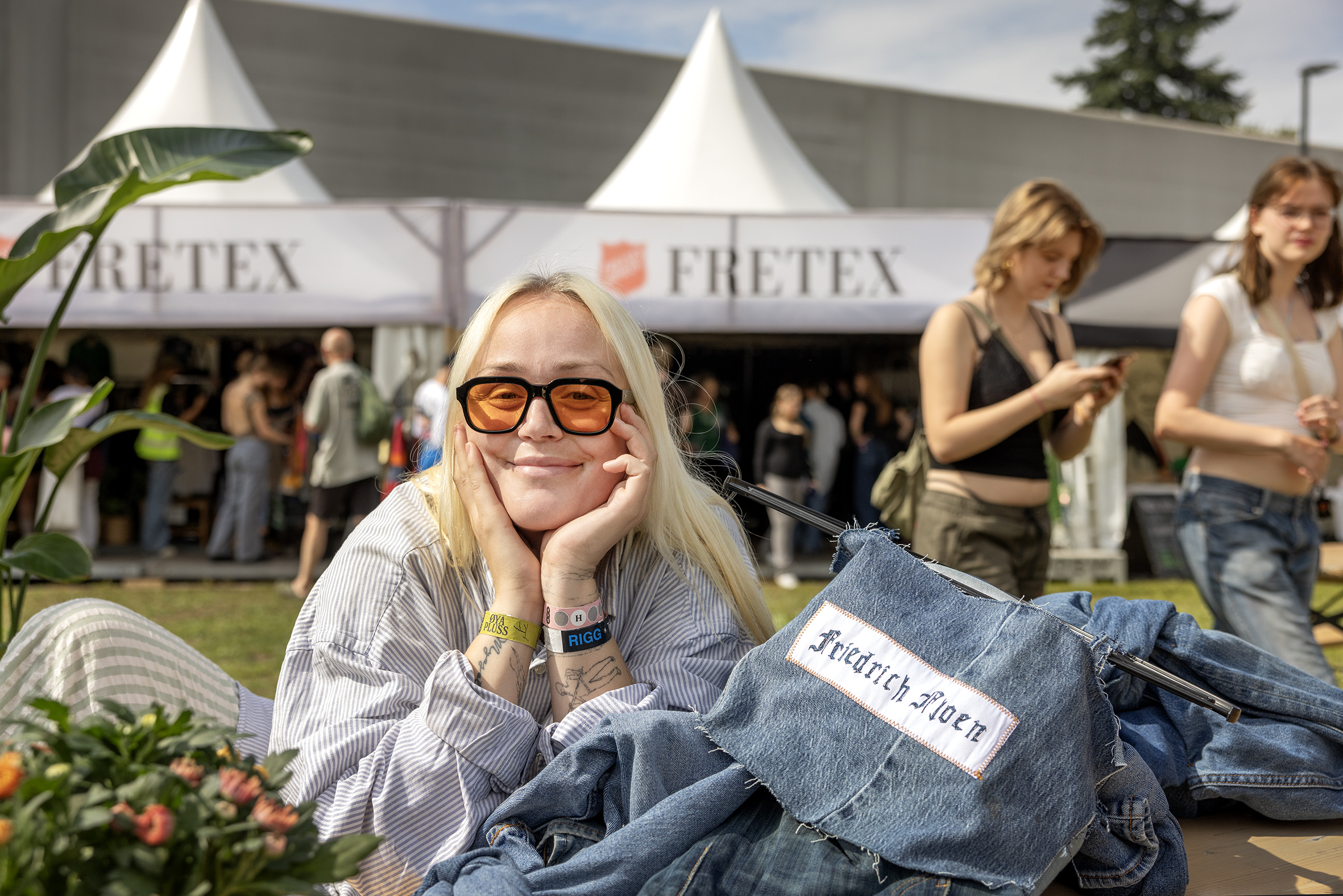Foto: Kvinne i 30-årene med blondt, langt hår og solbriller med gult glass kikker i kamera. Hun sitter med hodet i hendene og smiler. I bakgrunnen sees et telt fra Fretex.