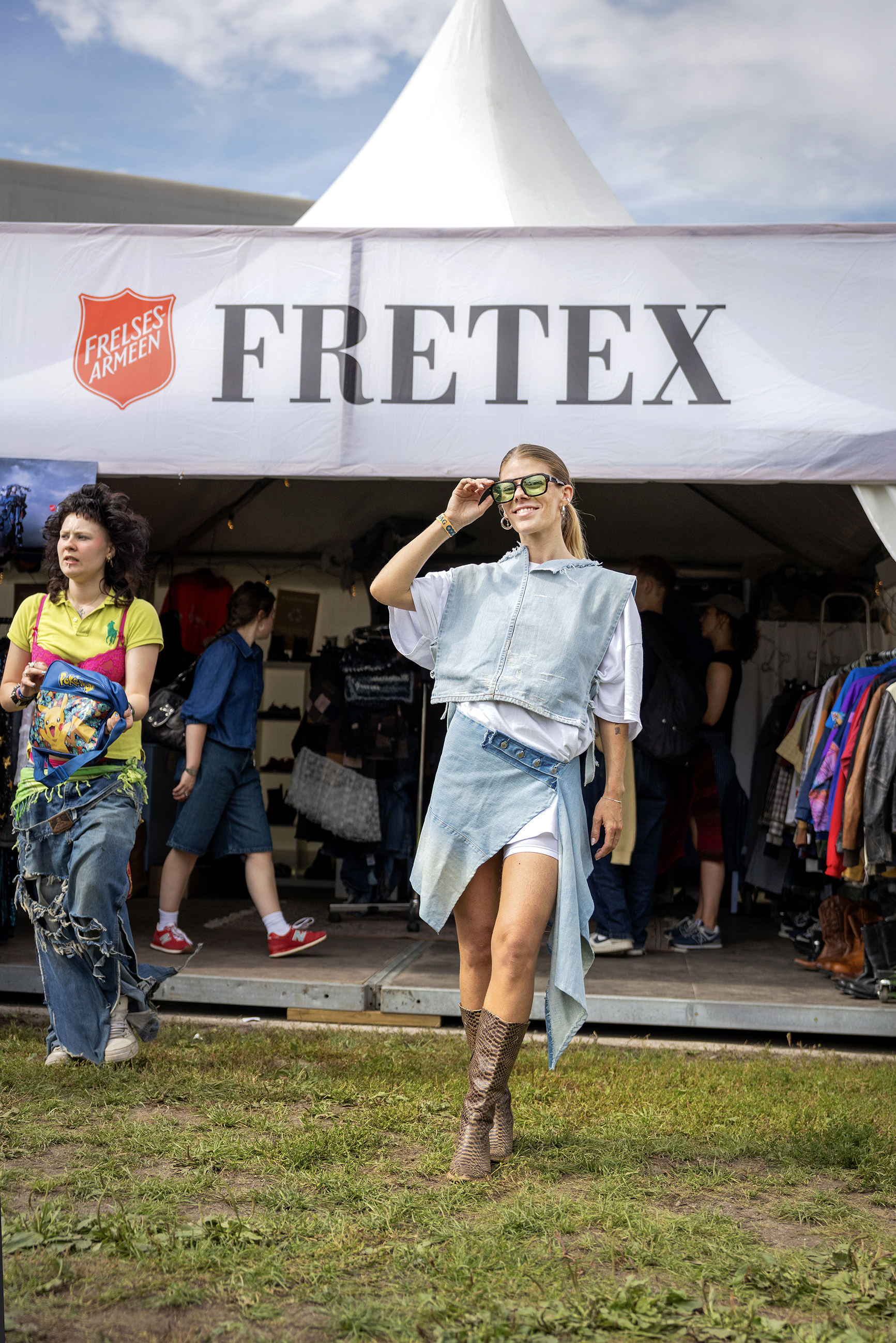 Foto: Ung kvinne med hestehale og solbriller poserer utenfor Fretex-teltet i et antrekk sydd av gamle jeans. Ut av butikken går en fargerik kunde med sjokkrosa BH oppå kanarigul T-skjorte.