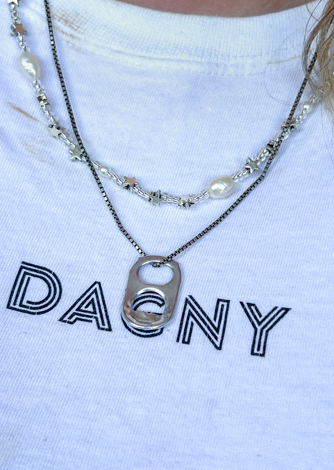 Foto: Hvit T-skjorte der det står Dagny med store bokstaver i en logoform. Over henger en bruskork i et kjedee sammen med noen andre smykker.
