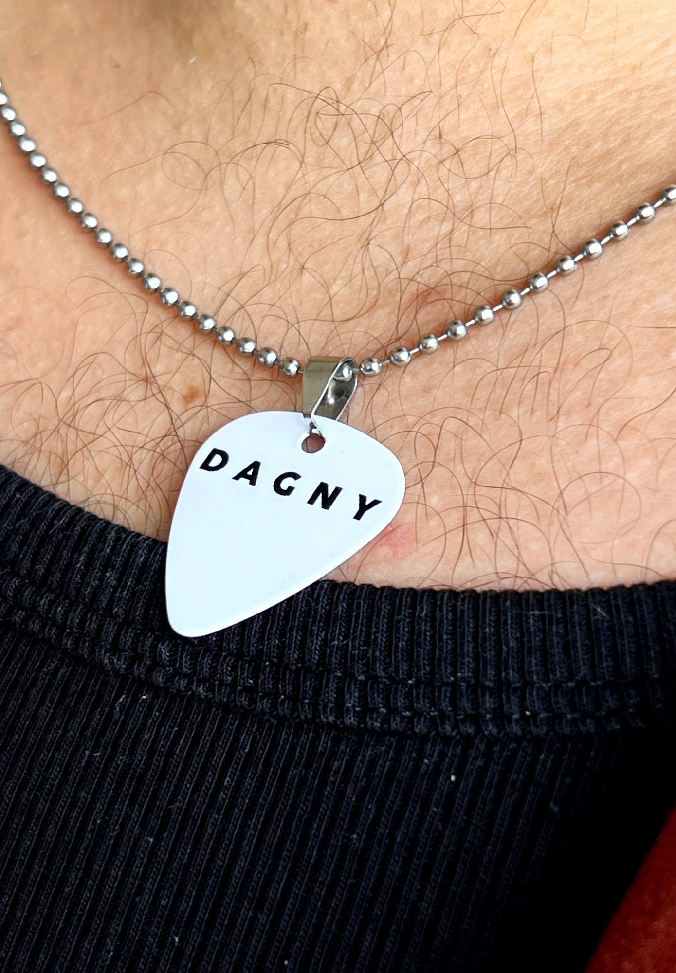 Foto: Brystet til en mann med hår. han har på seg en svart ribbet singlet og et hvitt smykke som ser mer ut som et plekter til en gitar. I toppen står det Dagny i store bokstaver.