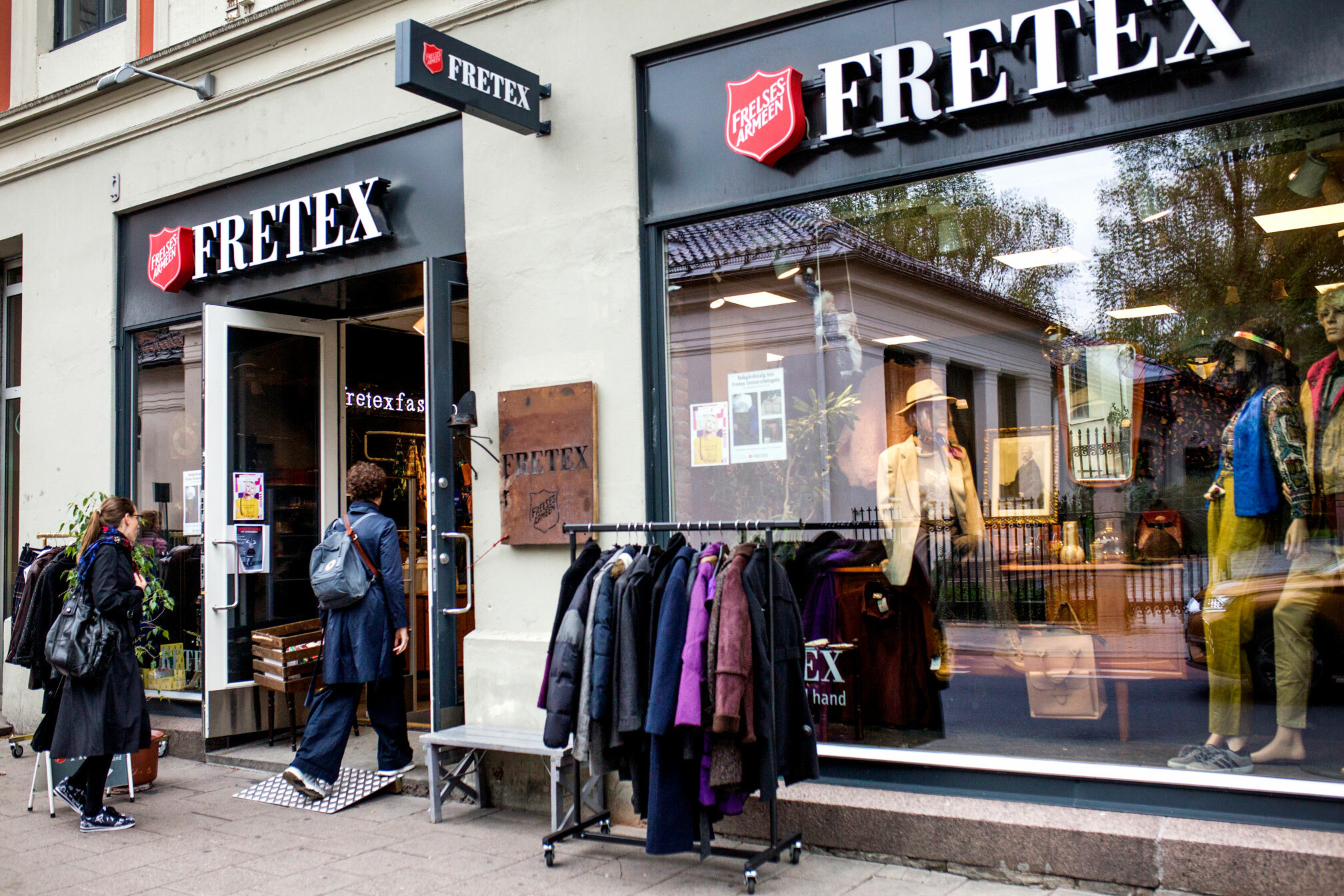Foto: Eksteriør av en Fretex-butikk. To mkvinner er på vei inn døra. Utenfor står det stativ med klær på.