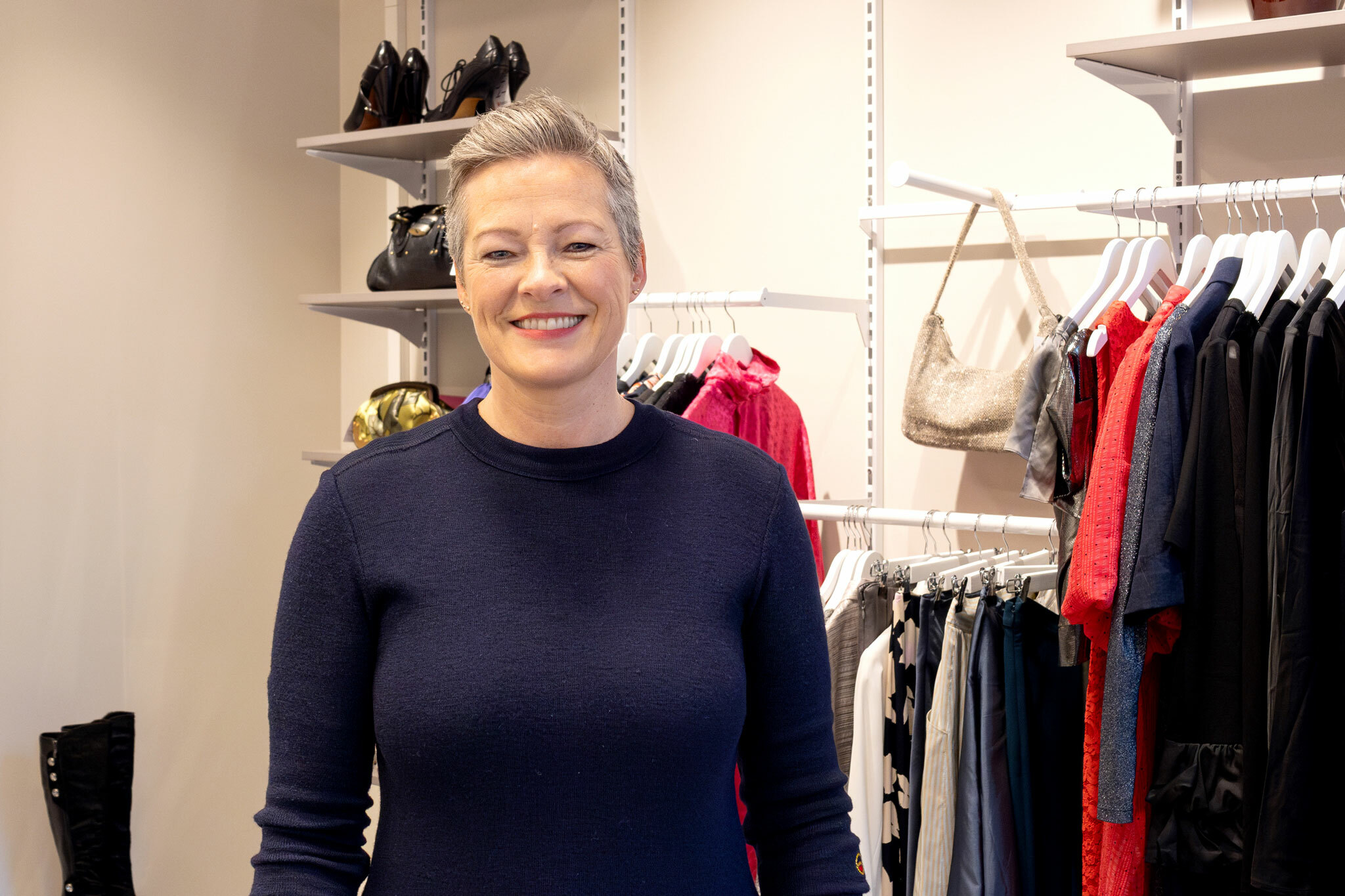 Foto: Kvinne i 50-årene med kort, grått hår og blå genser står i en bruktbutikk. Bak henne er det hyller med klær, sko og vesker.