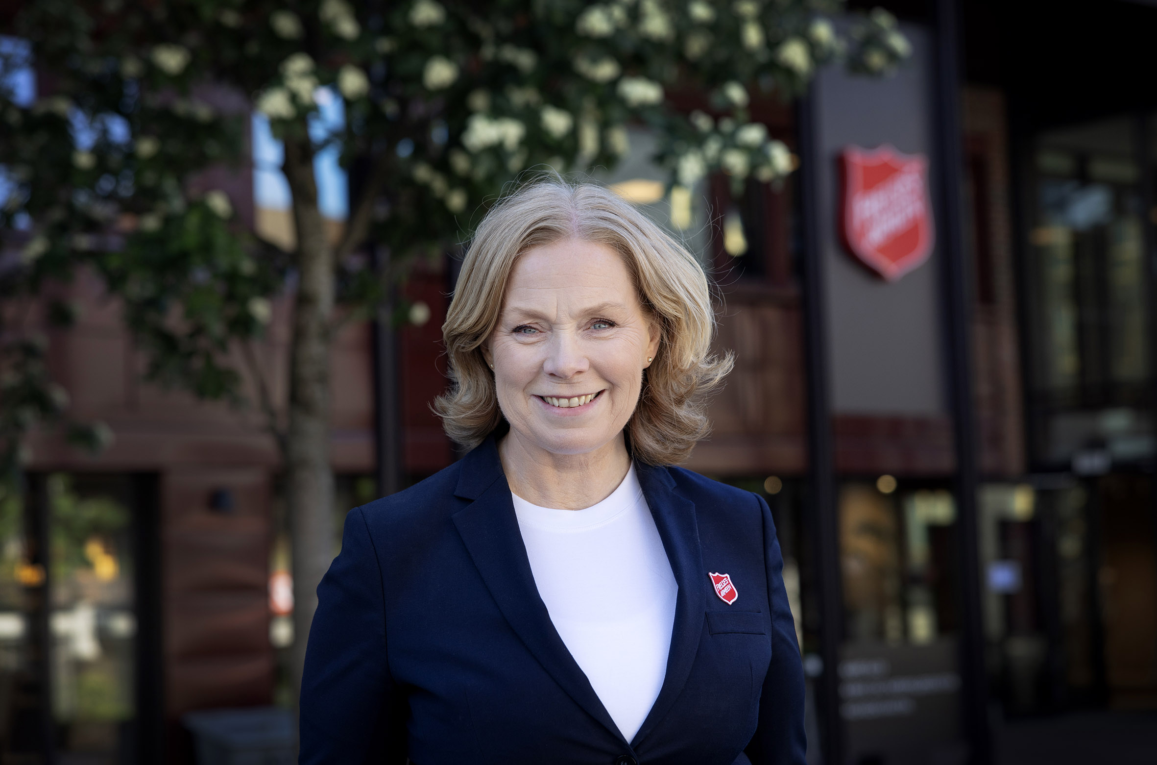 Foto: Dame i slutten av 40-årene står foran et stort bygg med logo fra Frelsesarmeen. Hun har gyllent hår og en mørkeblå dressjakke også med det røde skjoldet fra Frelsesarmeen på.