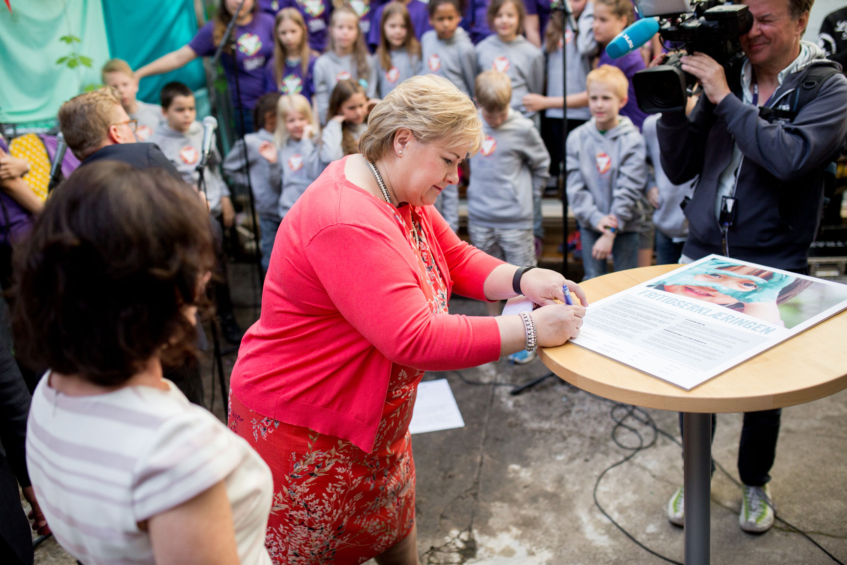 Foto: Blond kvinne i 50-årene med røde kjole og jakke utenpå signerer en kontrakt som ligger på et rundt ståbord. En mann fra NRk filmer med et stort kamera og det står en rekke barn i et kor i bakgrunnen.