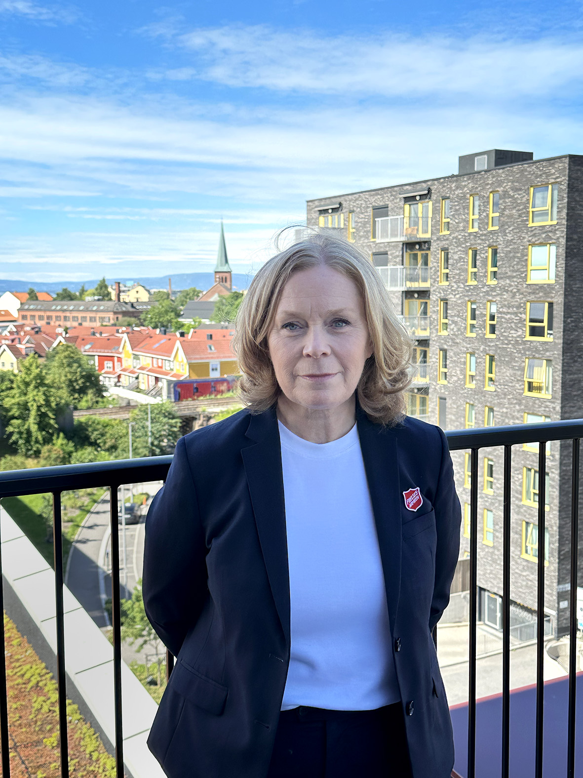 Foto: Kvinne med blondt hår står i en mørkeblå dressjakke med logo fra Frelsesarmeen. Hun står oppe på en terasse med utsikt over Oslo.