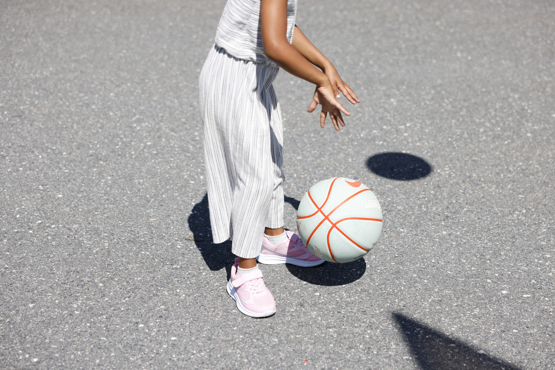 Foto: Jente med brun hud og rosa sko dabber en basketball ute på aspalt. Vi ser ikke hodet hennes, men hender som strekker seg ned mot ballen som er i lufta.