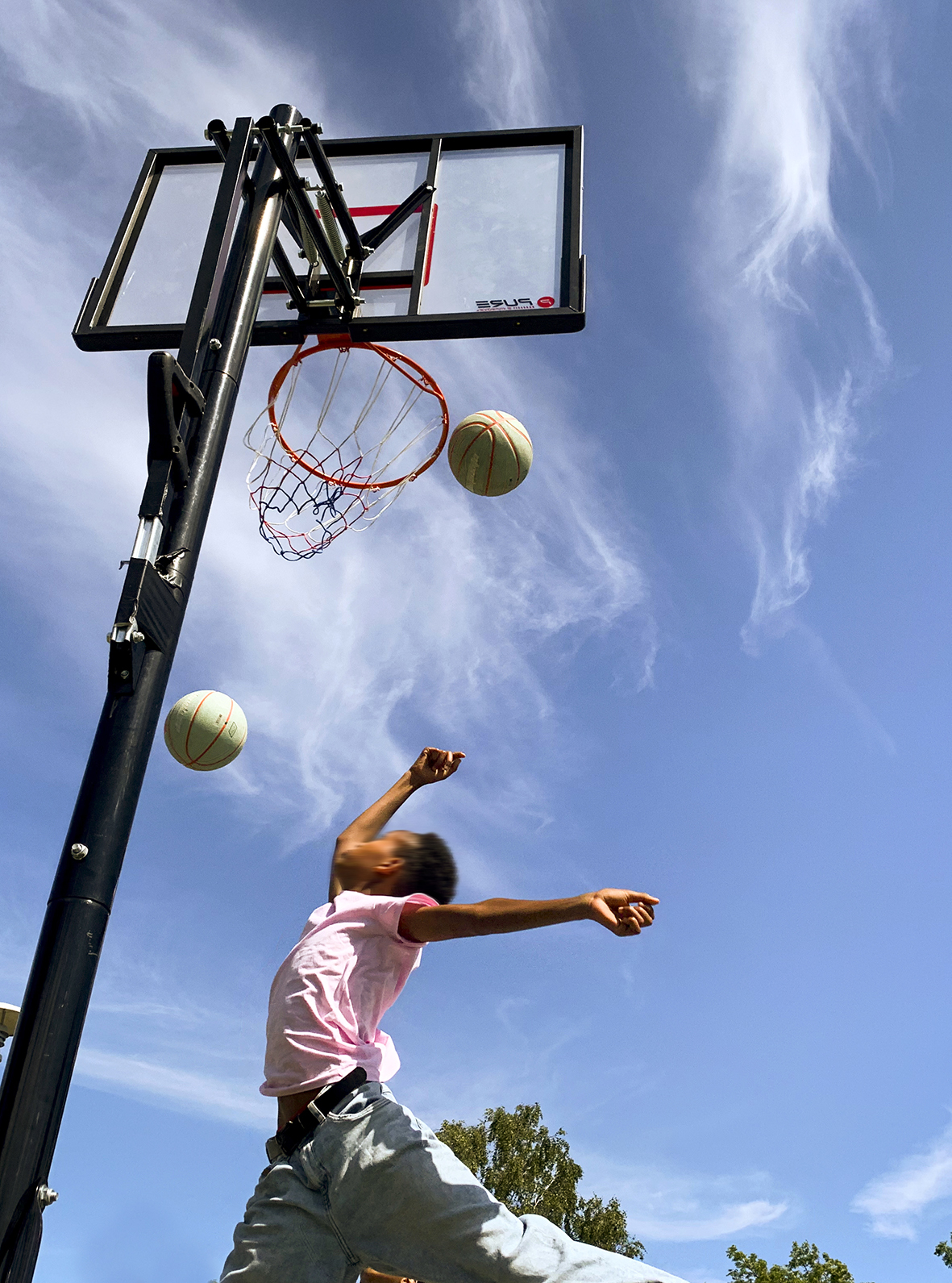 Foto: Barn med gyllen hud hopper høyt og kaster basketballen opp i kurven. Det er ute og blå himmel i bakgrunnen.