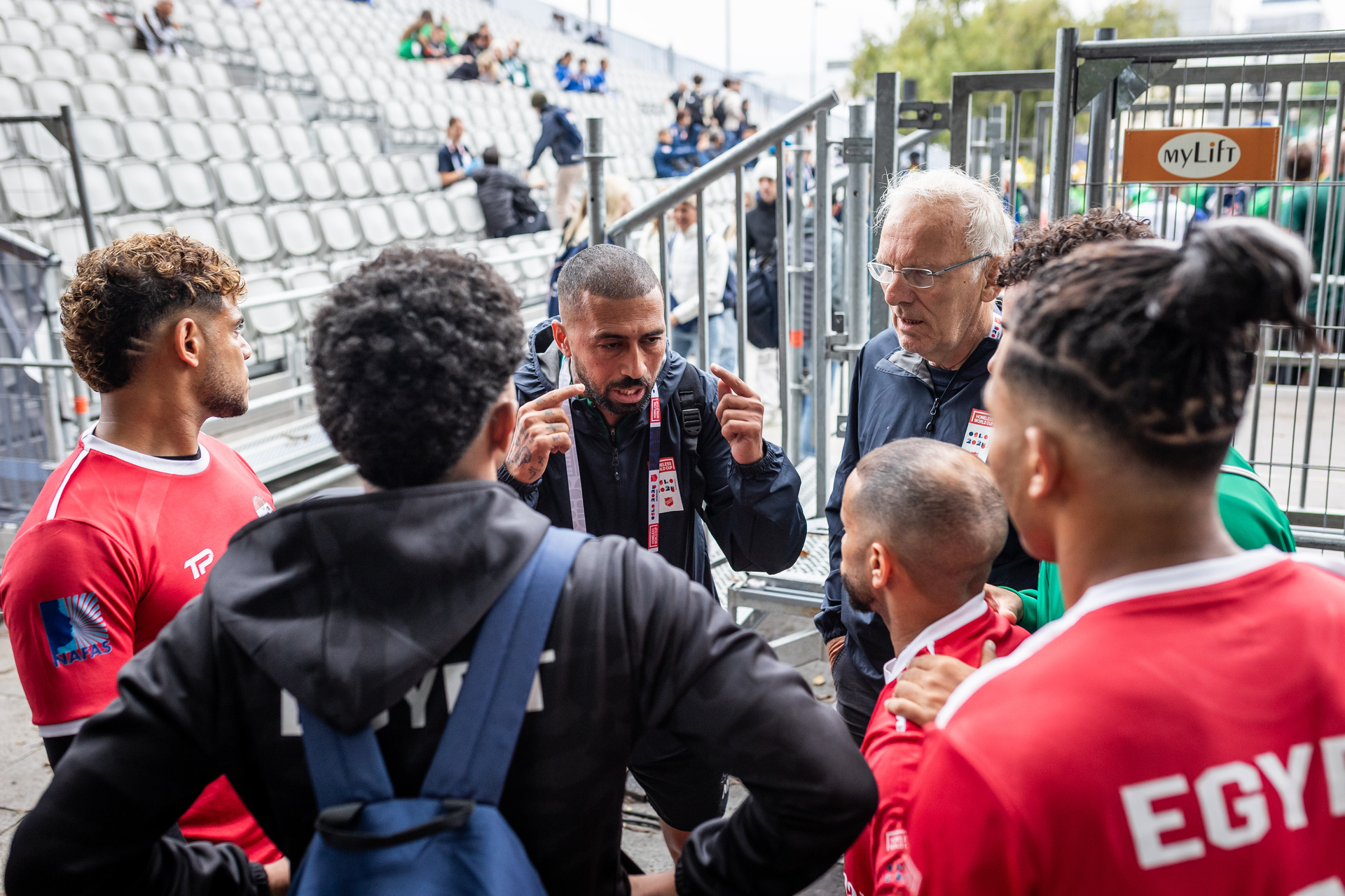 Foto: en gruppe egyptiske herrespillere står i en ring, de fleste med ryggen mot kamera. En spiller vendt mot kamera ser ut til å være i en samtale med en spiller, han peker mot hodet sitt. Til høyre i gruppa står Terje Thodesen med grått hår, briller og blå jakke.