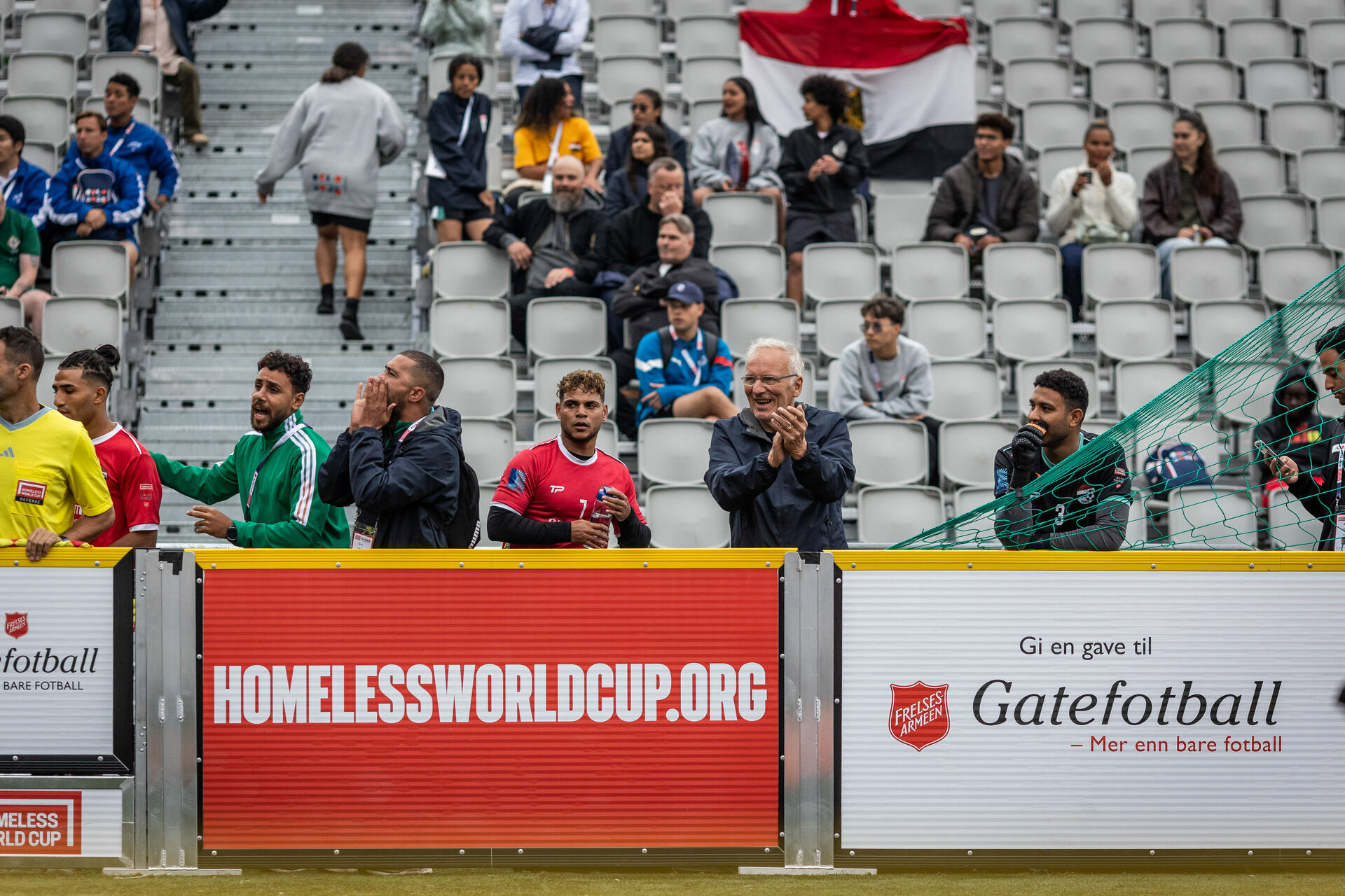 Foto: Terje Thodesen står rett utenfor banen og klapper under en kamp. Andre spillere som ikke er på banen da står ved siden av og roper og klapper. I bakgrunnen sees tribunen med andre publikummere.