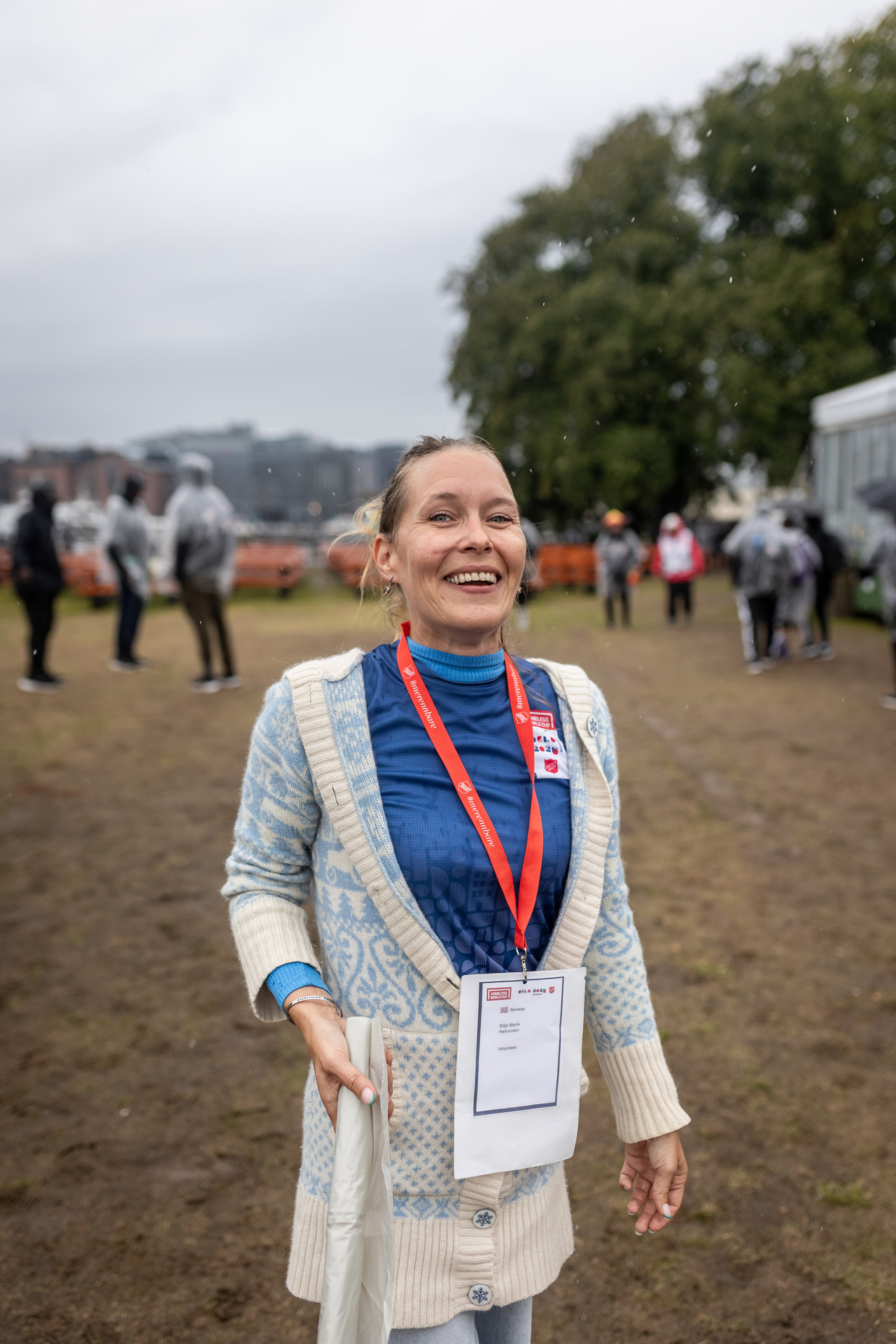 Foto: Silje Marie står og smiler mot kamera. Hun har på seg blå t-skjorte med HWC-logo og en hvit og blå strikkejakke over. I hånden holder hun en rull med søppelposer. I bakgrunnen skimtes fotballspillere i regnponcho.