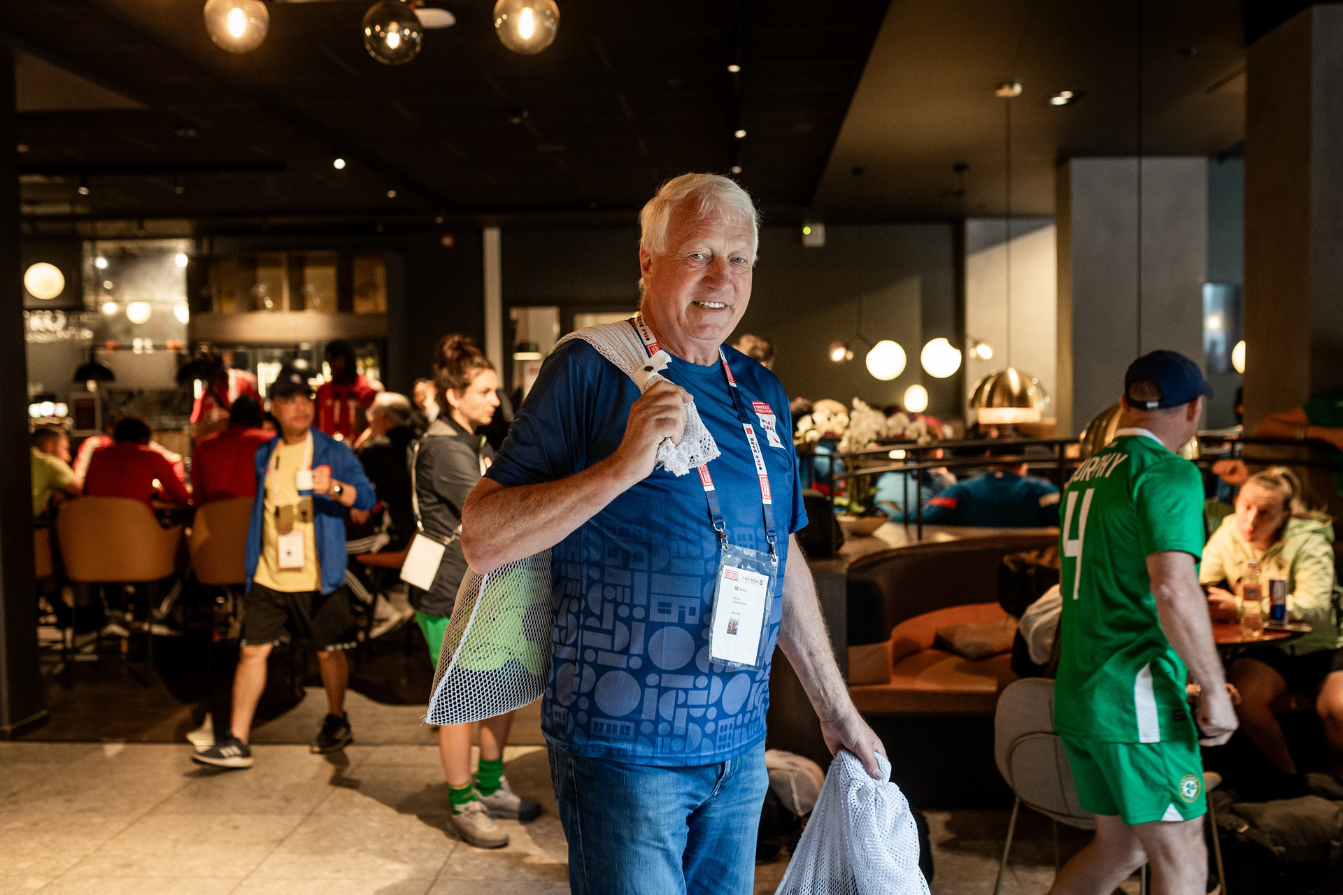 oto: En eldre mann står og holderen hvit sekk i hånda, og har en annen sekk slengt over skulderen. Han har på seg blå hwc-t-skjorte. Bak ham sees folk som går rundt i resepsjonen på hotellet der han er frivillig,