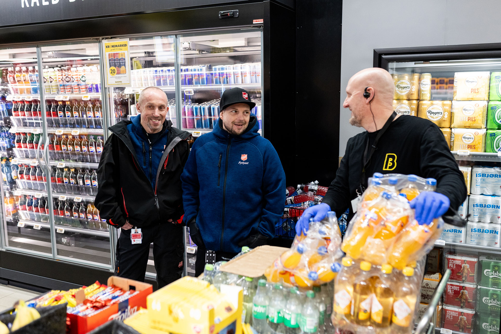 Foto: Skallet mann med svart genser med gul B, logoen for Bunnpris, står i en butikk. Han har en stabel med brusflasker i kasser foran seg og snur hodet for å snakke med to menn med logo fra Frelsesarmeen på klær og nøkkelkort. En av dem har caps.
