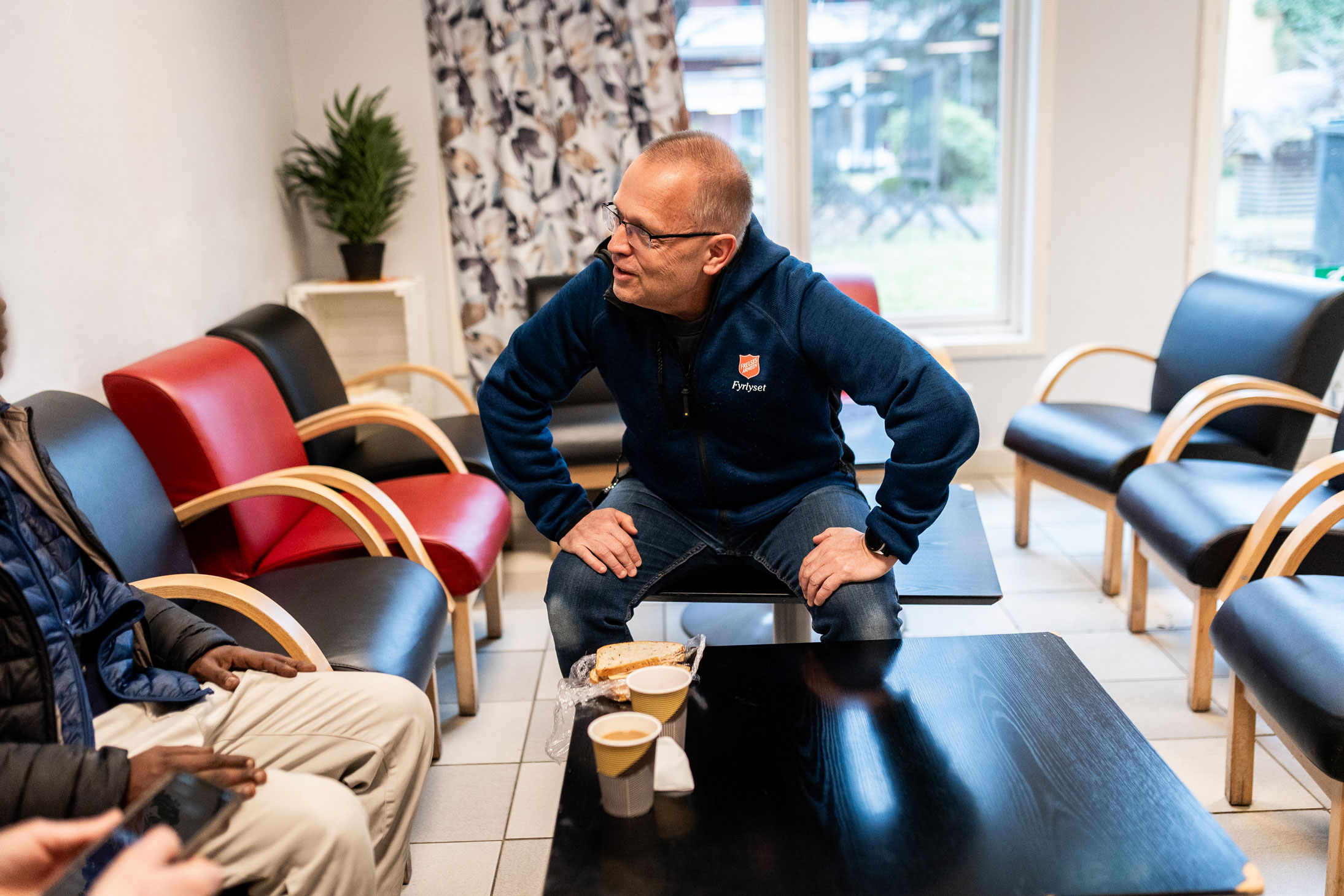 Foto: Mann i 50-årene med briller og kort hår sitter i i en lenestol ved et sofabord og snakker med et par personer vi ikke ser. Mannen har på seg blå hettegenser fra Frelsesarmeen og har på seg et rødt skjold på brystet. Han lener seg framover og holder begge hendene på lårene sine.