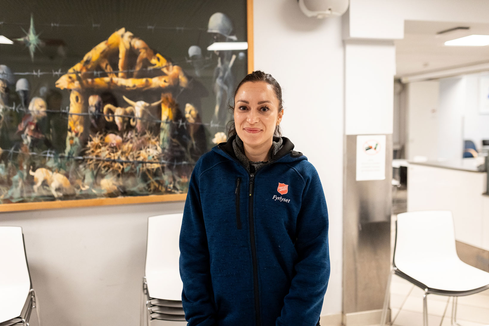 Foto: Kvinne i 30-årene med mørkt hår har på seg blå flisjakke med logo fra Frelsesarmeen. Under står det Fyrlyset. Hun har brune øyne og smiler gjennom dem mer enn munnen. Hun står langs en vegg, foran et bilde på veggen og hvite stoler i stabler.