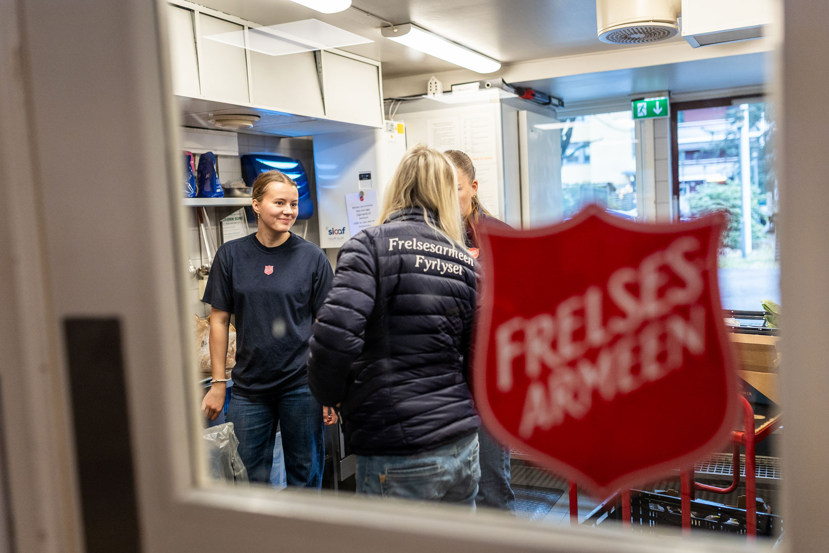 Foto: Gjennom en hvit dør med vindu ser vi inn til et rom men tre damer. På ryggen til en av det står det Frelsesarmeen Fyrlyset med hvite bokstaver på en mørkeblå boblejakke. På glasset i vinduet er det et stort skjolde der det står Frelsesarmeen.
